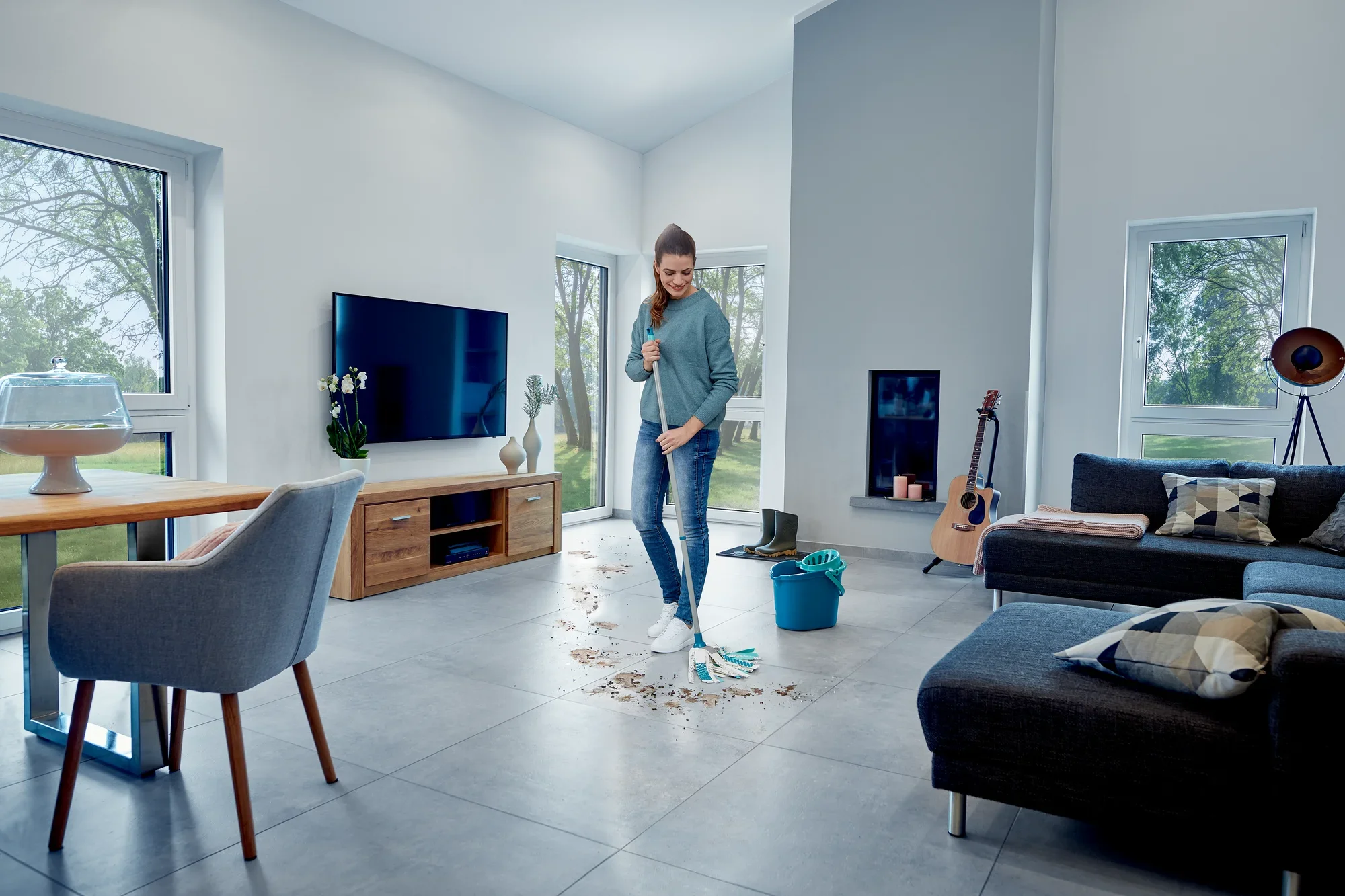 A woman is cleaning the floor of a modern living room with the Power Mop 3in1. The room is bright and stylishly furnished, with a large window and a comfortable sofa.