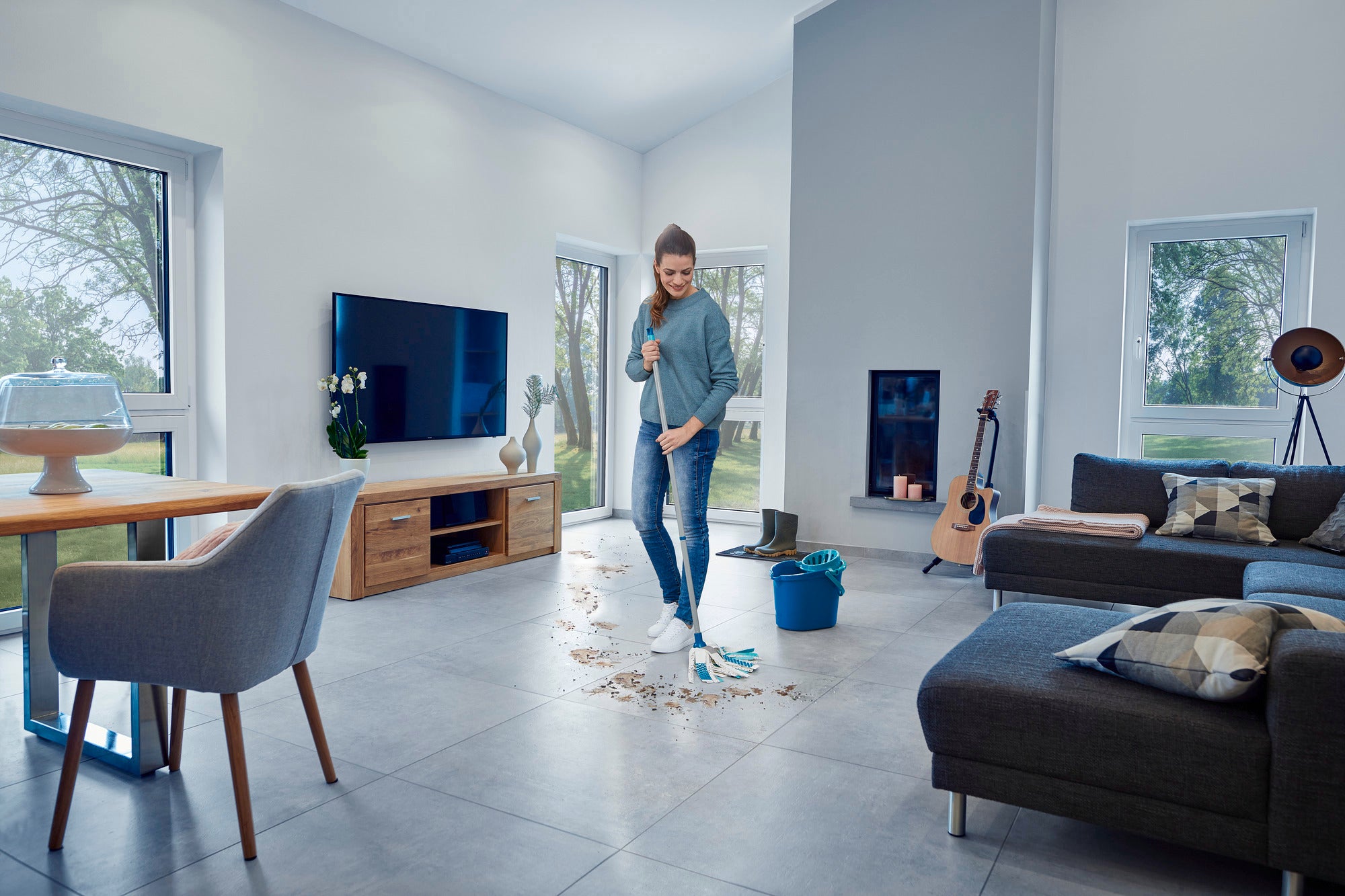 Une femme nettoie le sol d'un salon moderne avec le Power Mop 3in1. La pièce est lumineuse et élégamment meublée, avec une grande fenêtre et un canapé confortable.