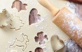 Weihnachtsbäckerei zum Verschenken und selber essen Ein Nudelholz und ausgestochene Plaetzchen samt Formen liegen auf einem ausgerollten Teig auf einer bemehlten Arbeitsflaeche.
