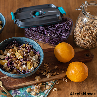 Eine Schuessel mit Rotkohlsalat, Orangenstuecken und Walnuessen auf einem Holzbrett, daneben frische Orangen und ein Glas Walnuesse.