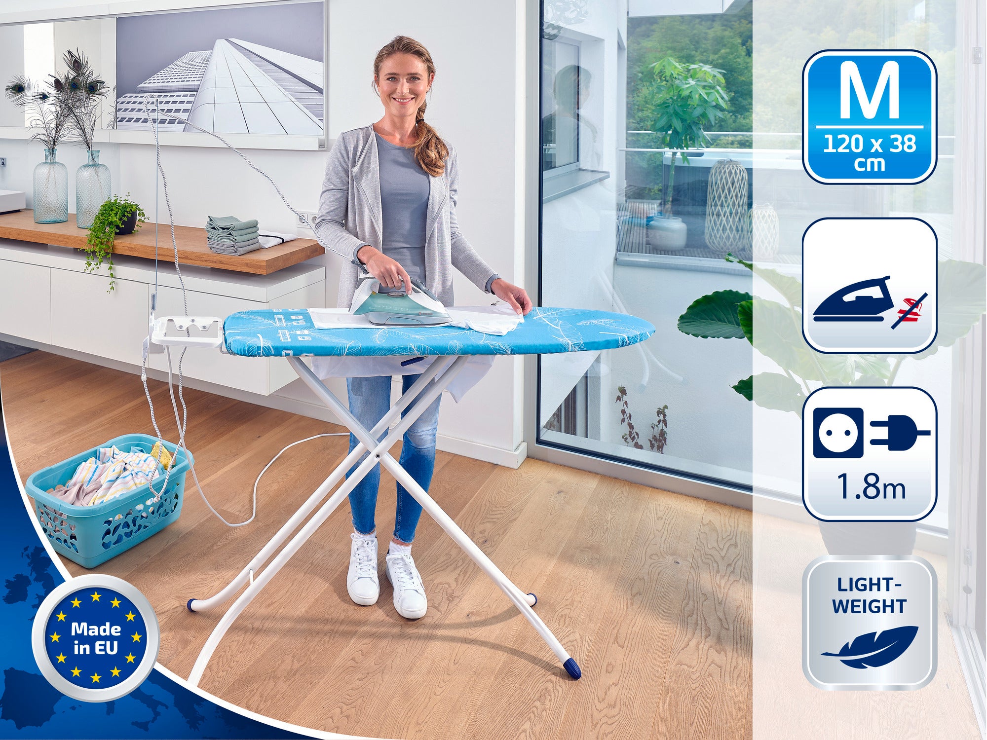 Una mujer plancha sobre una mesa de planchar azul en una cocina moderna. La mesa mide 120 x 38 cm, es ligera y tiene un cable de 1,8 m de longitud. Fabricada en la UE.