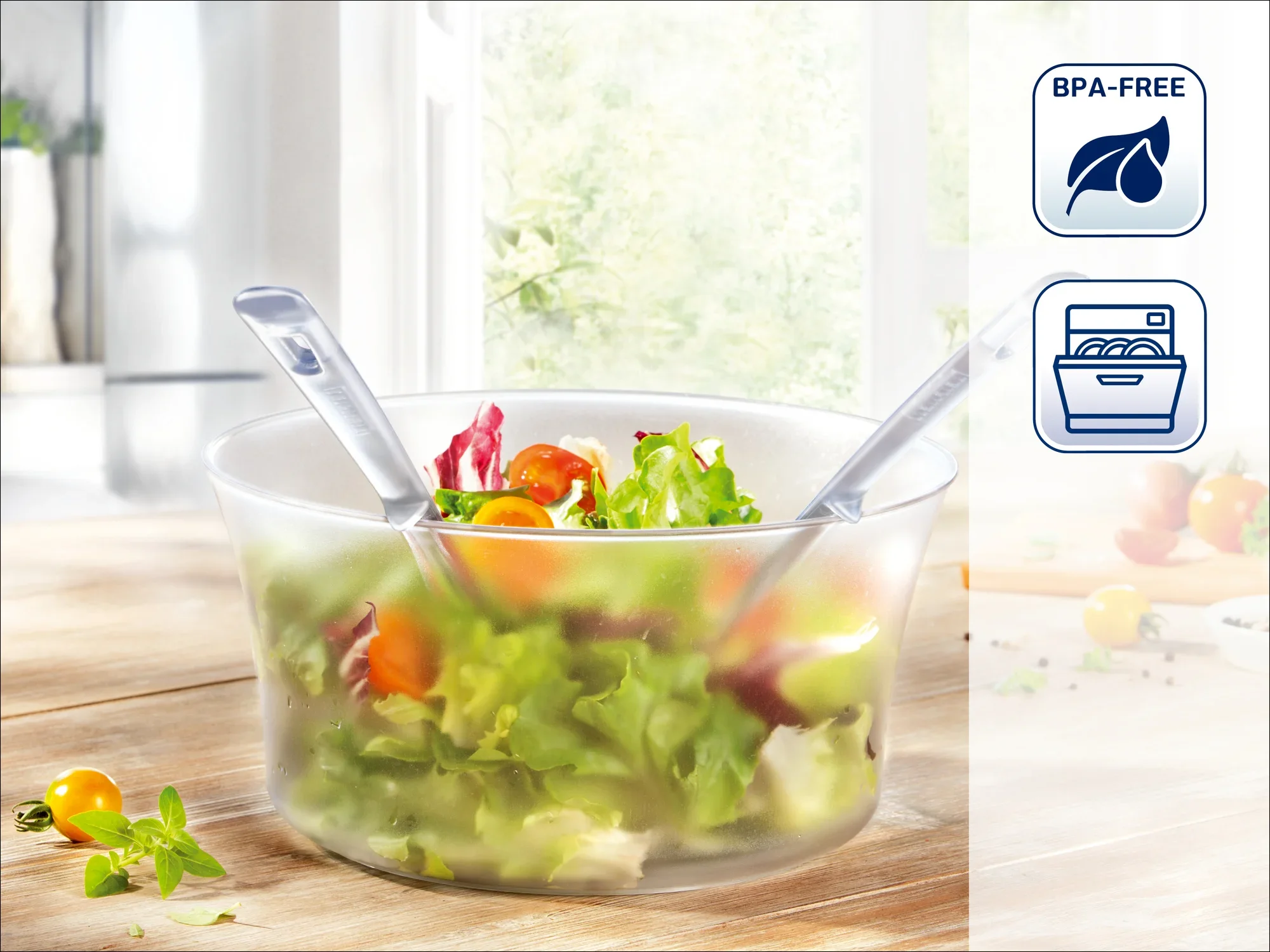 Una ensaladera transparente con lechuga fresca y dos cucharas para servir. Al fondo, una cocina luminosa. Los símbolos indican que no contiene BPA y que es apto para el lavavajillas.