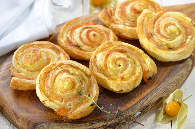 Sechs goldbraune Blaetterteigschnecken mit Frischkaese und Lachs auf einem rustikalen Holzbrett, garniert mit einem Thymianzweig und einer Physalis.