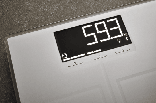 Digitale Waage auf dunklem Boden zeigt 59,3 kg an, mit Symbolen fuer Batteriestatus, Geschlecht und Bluetooth-Verbindung auf dem Display.