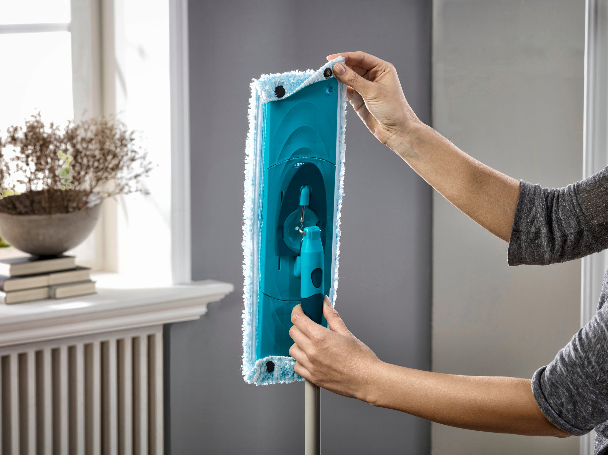 Eine Person zeigt, wie der CLEAN TWIST Moppkopf gewechselt wird. Der Mopp ist blau und wird in einem hellen Raum mit einem Fenster im Hintergrund gehalten. Ideal für eine gründliche Bodenreinigung.