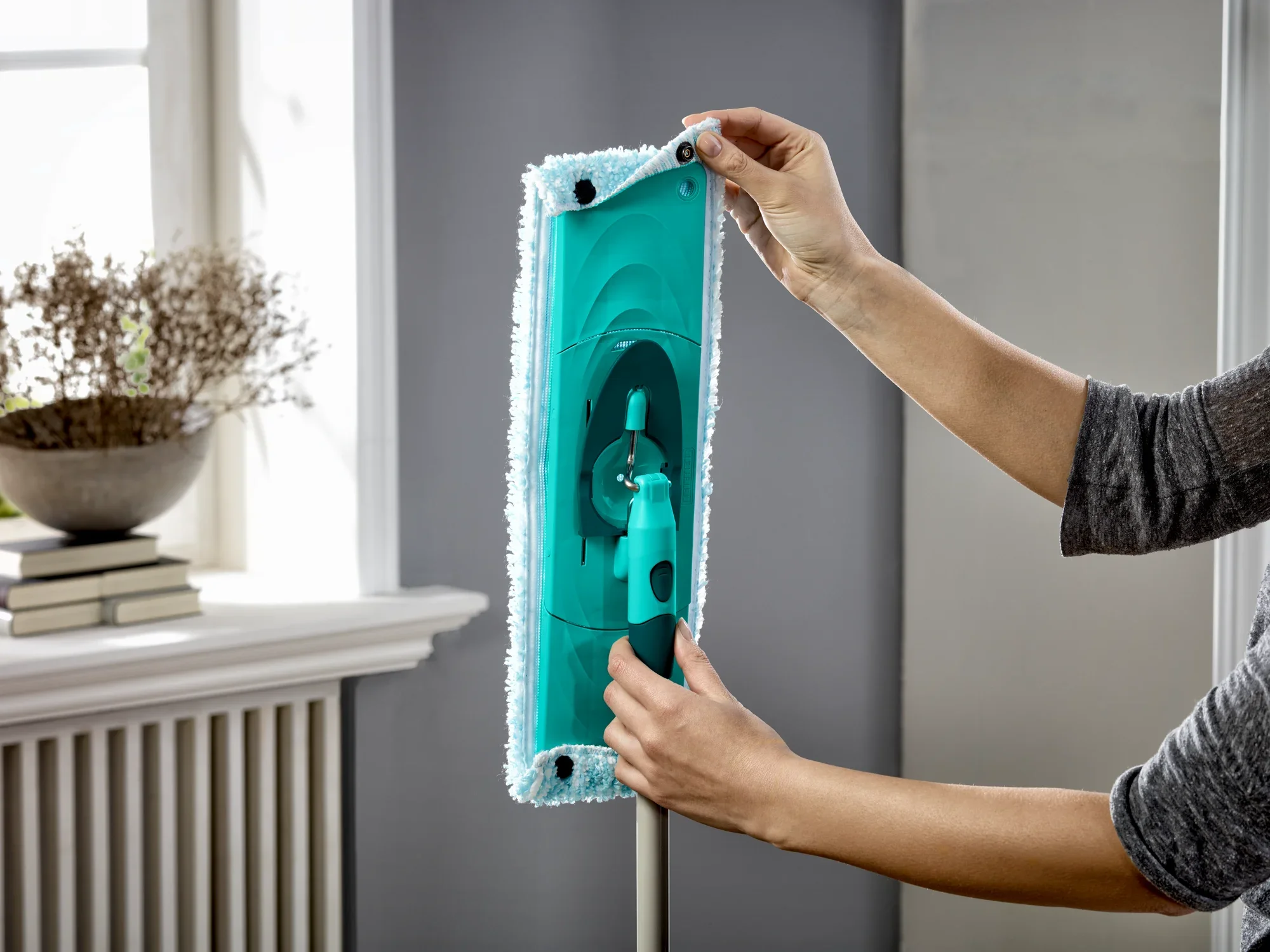 Eine Person zeigt, wie der CLEAN TWIST Moppkopf gewechselt wird. Der Mopp ist blau und wird in einem hellen Raum mit einem Fenster im Hintergrund gehalten. Ideal für eine gründliche Bodenreinigung.