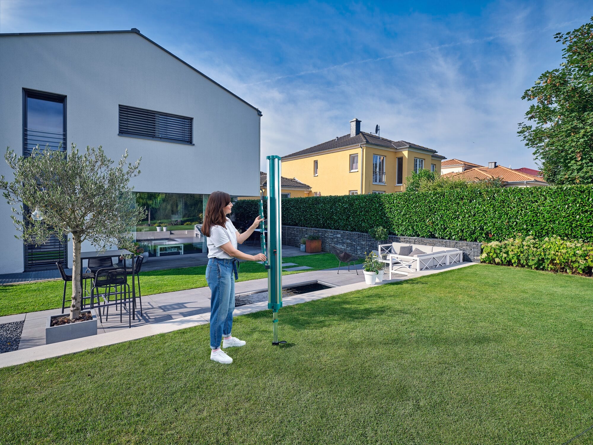 Eine Frau steht in einem gepflegten Garten und befestigt den Easy Clip Kleinteilehalter an einem Linomatic Wäscheschirm. Im Hintergrund sind moderne Häuser und eine Hecke zu sehen.