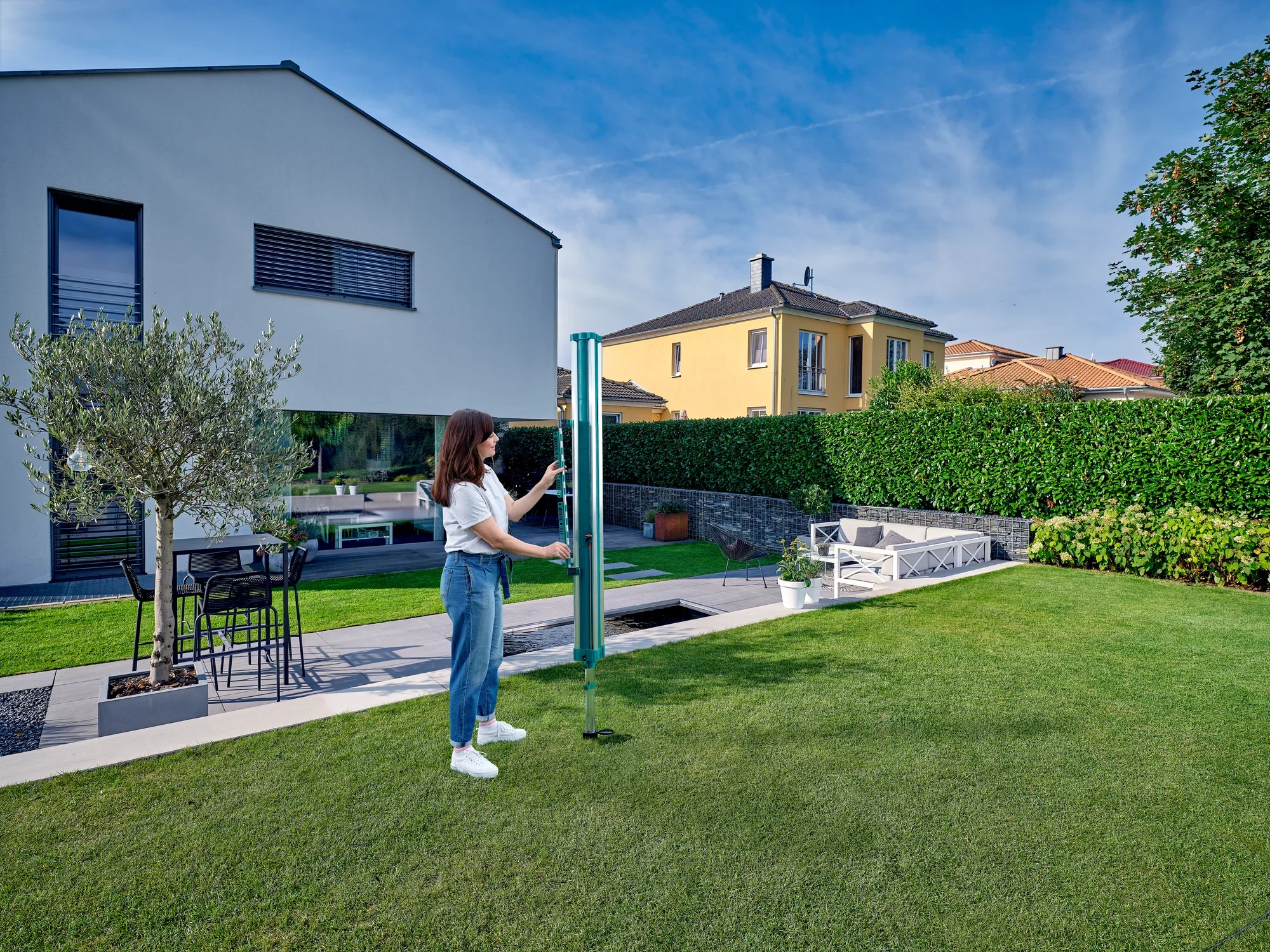 Kobieta stoi w zadbanym ogrodzie i mocuje uchwyt na drobne części Easy Clip do parasola na pranie Linomatic. W tle widać nowoczesne domy i żywopłot.