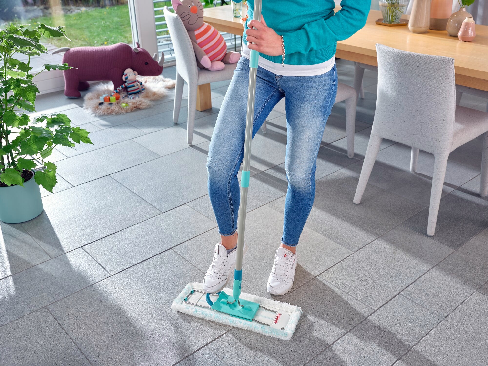 Eine Person reinigt einen hellgrauen Fliesenboden mit der Wischtuchpresse Profi XL. Im Hintergrund sind ein Esstisch, Stühle und ein Spielzeug zu sehen. Die Szene wirkt hell und einladend.