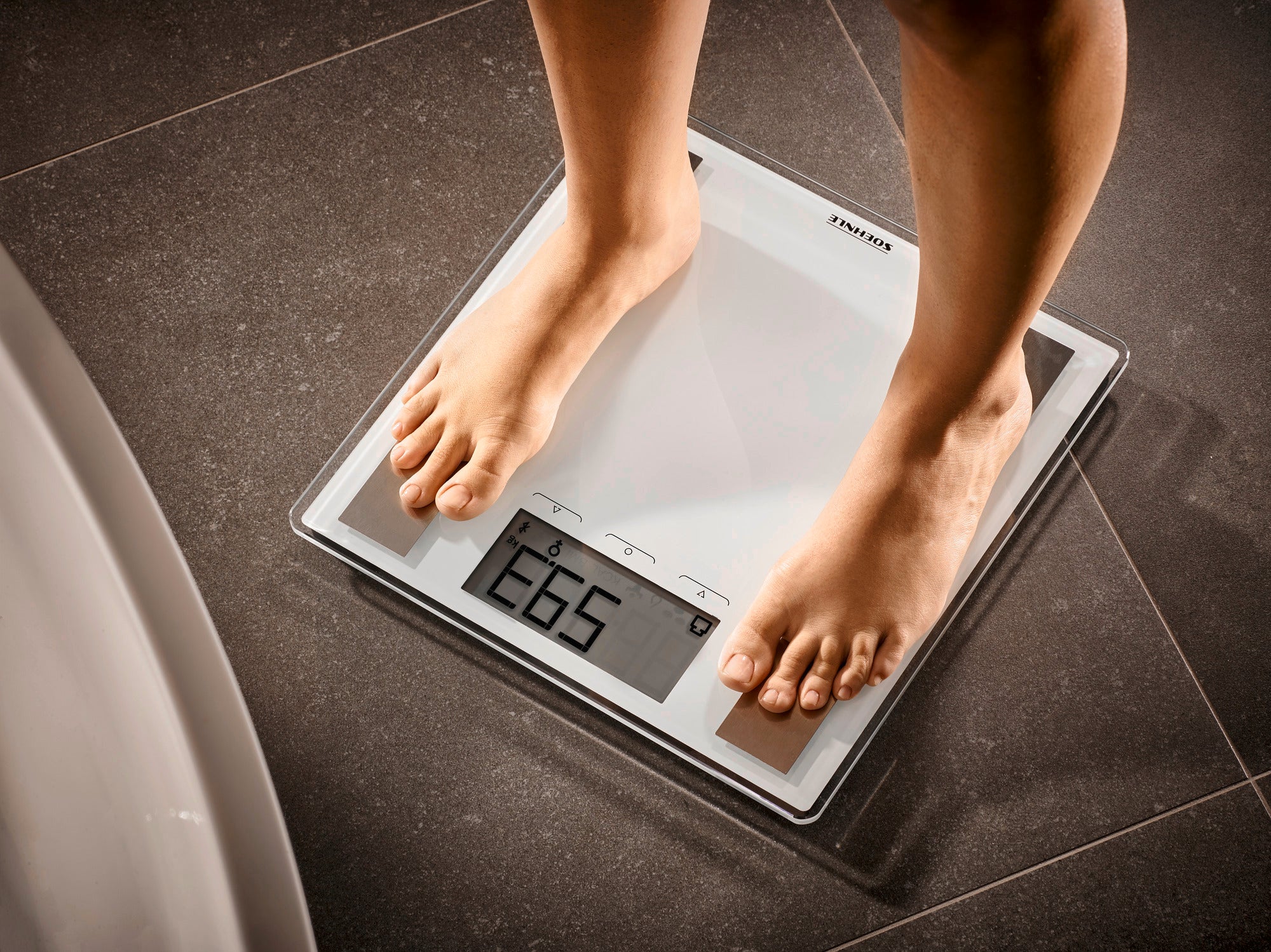 A person is standing on the Shape Sense Connect 100 digital bathroom scale. The scale shows a weight of 68.5 kg. It has a modern design and a clear display.