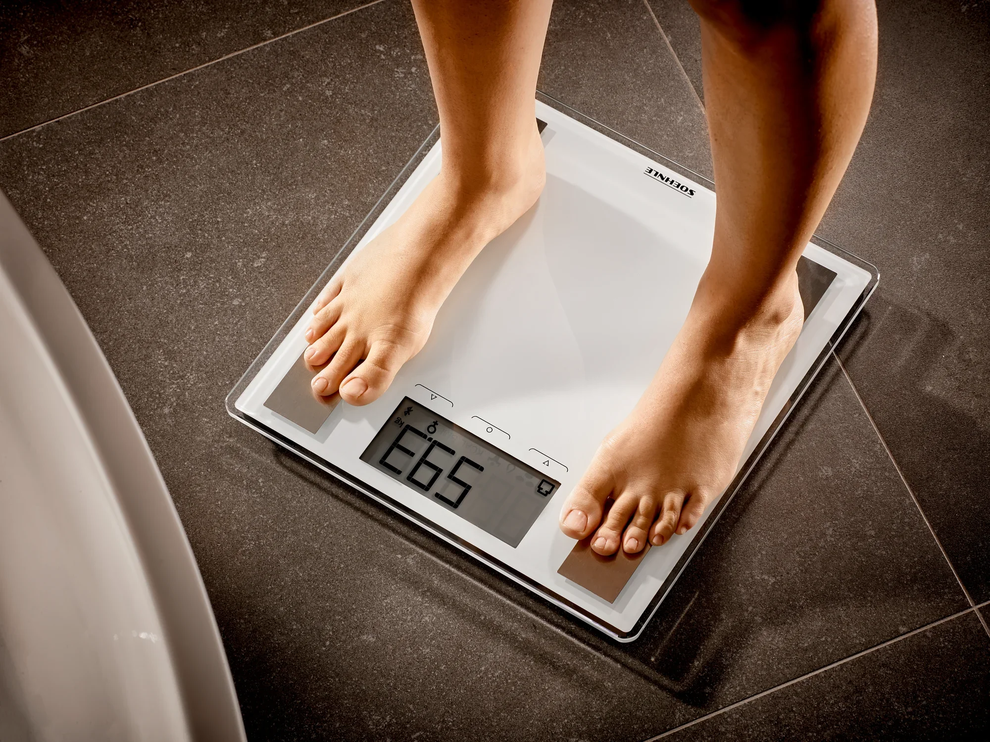 Een persoon staat op de Shape Sense Connect 100 digitale badkamerweegschaal, die een gewicht van 68,5 kg weergeeft. De weegschaal heeft een modern design en een duidelijk display.