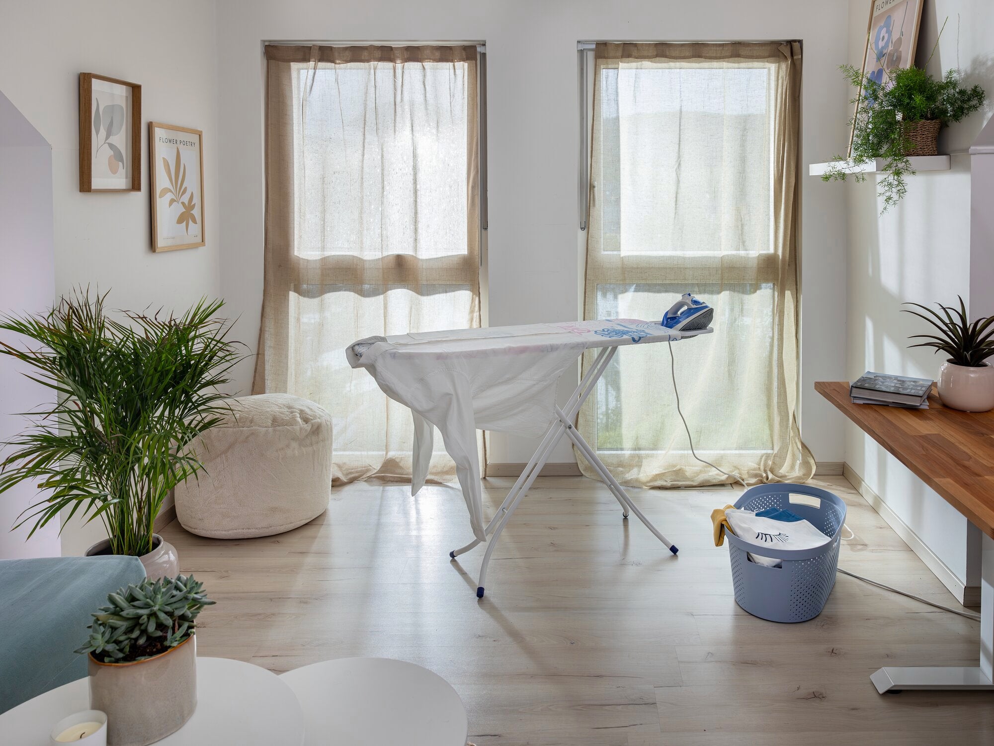 Une table à repasser Classic M Basic se trouve dans une pièce lumineuse avec deux grandes fenêtres. La table est équipée d'un fer à repasser. Des plantes et des décorations complètent l'ambiance chaleureuse.
