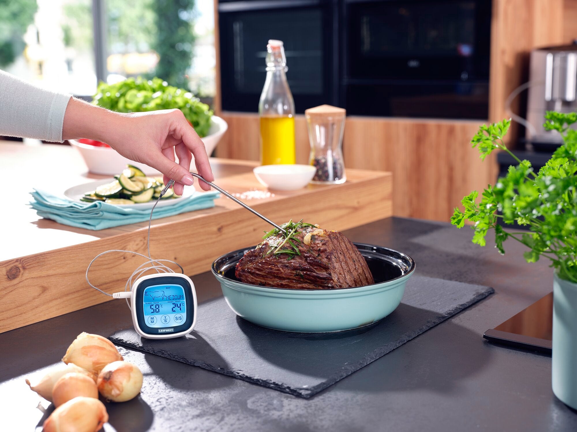 Eine Hand benutzt ein Braten- und BBQ-Thermometer, um die Temperatur eines Bratens in einem Topf zu messen. Im Hintergrund sind Küchenutensilien und frische Zutaten zu sehen.