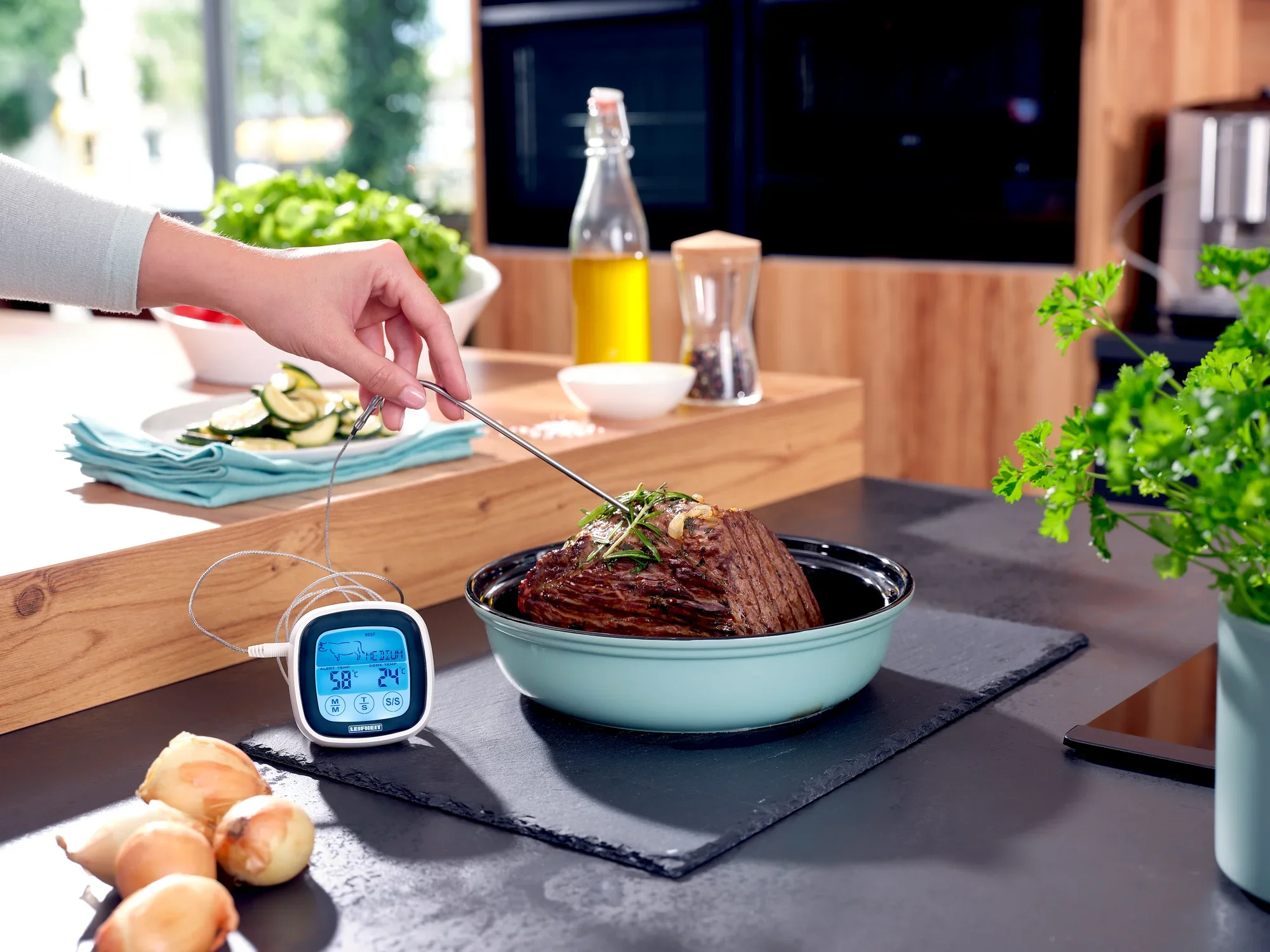 Jedna ręka używa termometru do pieczenia i grillowania, aby zmierzyć temperaturę pieczeni w garnku. W tle widoczne są przybory kuchenne i świeże składniki.