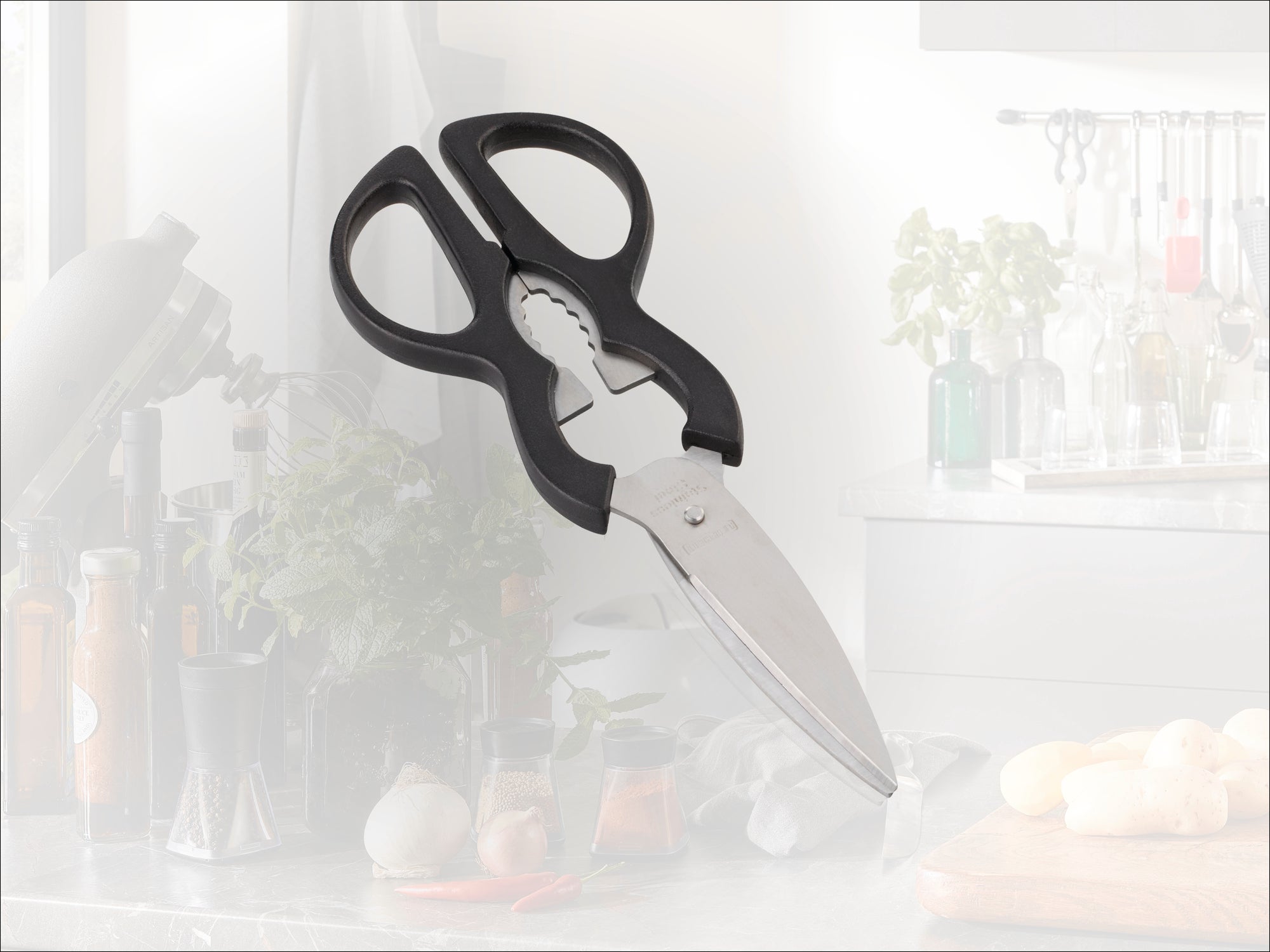Ciseaux de cuisine avec poignées noires et décapsuleur intégré, posés sur un plan de travail. Des herbes aromatiques et des ustensiles de cuisine sont visibles à l'arrière-plan.