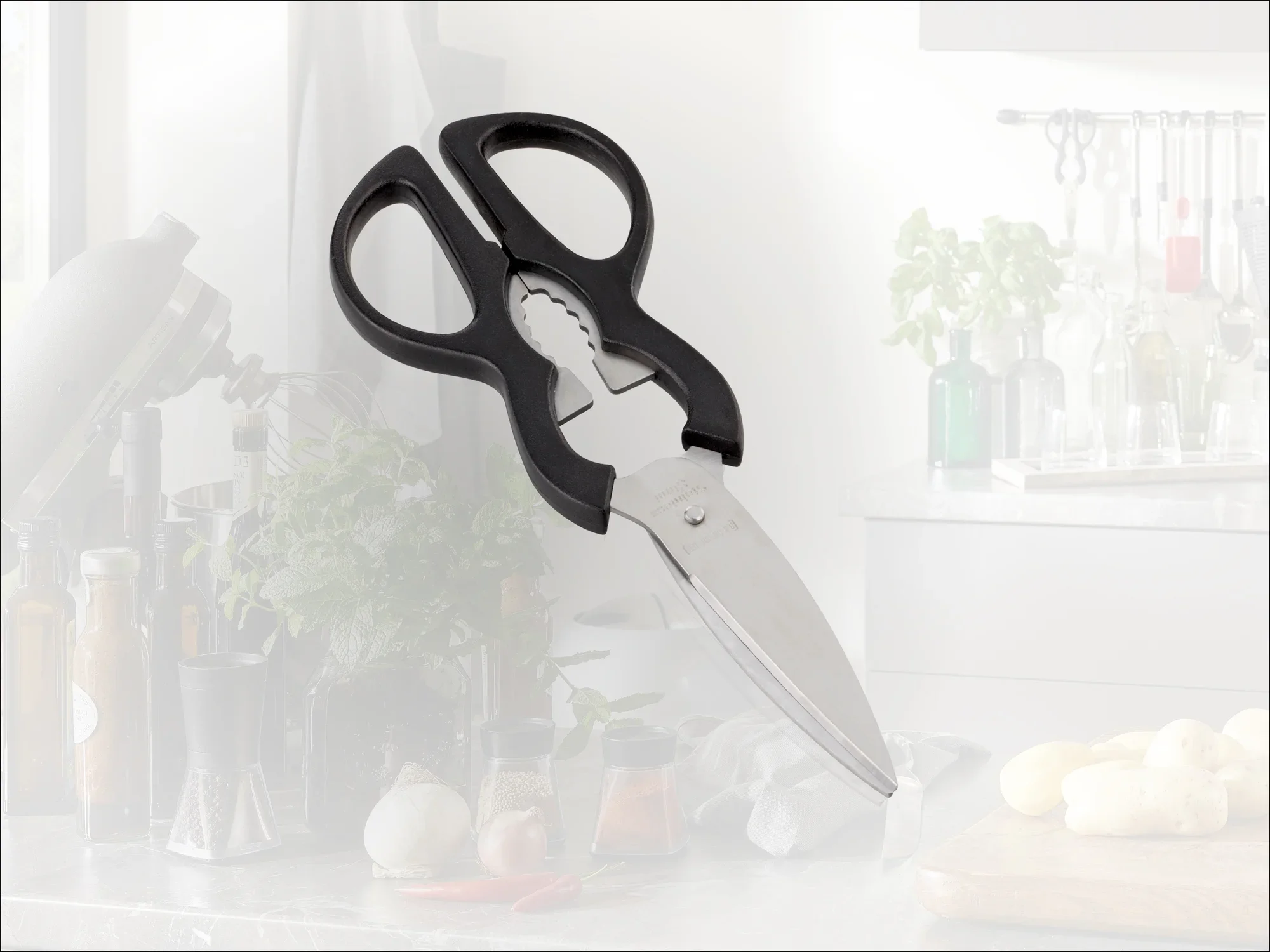 Nożyczki kuchenne z czarnym uchwytem i zintegrowanym otwieraczem do butelek, leżące na blacie kuchennym. W tle widoczne są zioła i przybory kuchenne.