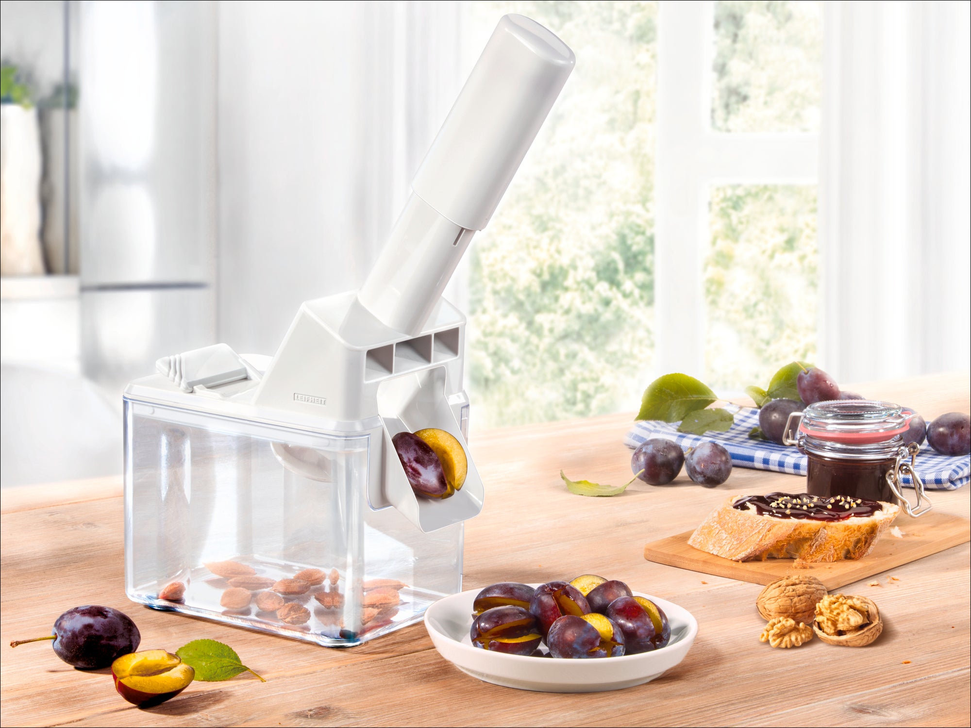 Un dénoyauteur de prunes blanc est posé sur une table en bois. Il enlève les noyaux des prunes qui tombent dans une boîte transparente. À côté se trouvent des prunes fraîches et un pot de confiture.