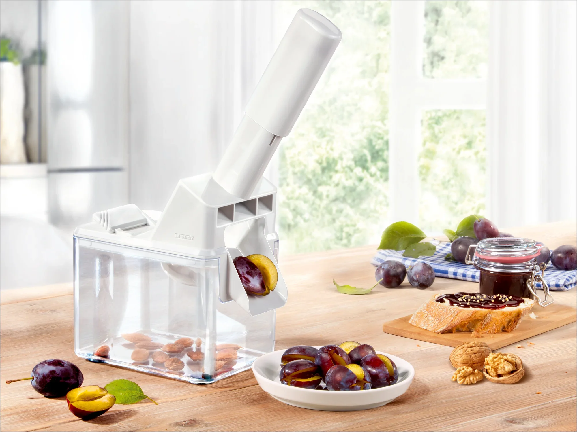 Biała drylownica do śliwek stoi na drewnianym stole. Usuwa pestki ze śliwek, które wpadają do przezroczystego pudełka. Obok leżą świeże śliwki i słoik dżemu.