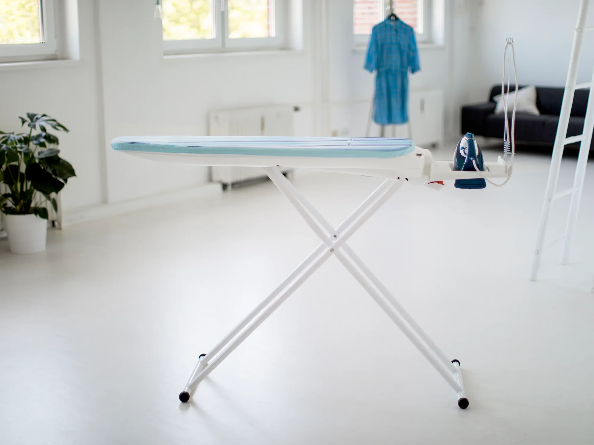 Table à repasser Air Active Express M dans une pièce lumineuse. La table est blanche avec une surface de repassage bleue, à côté se trouve un fer à repasser vapeur. Une robe bleue est suspendue à l'arrière-plan.