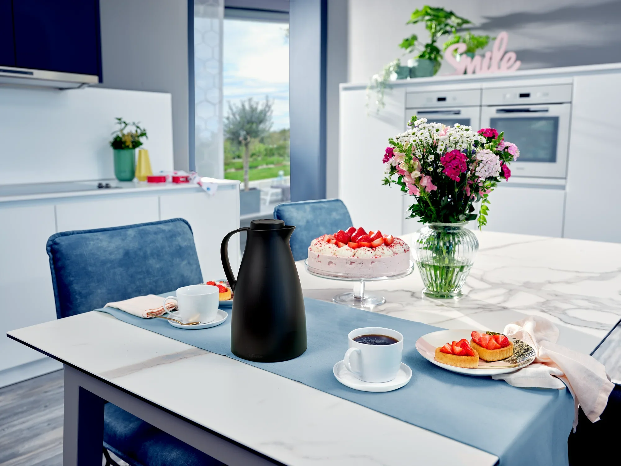 Una elegante cocina con una jarra térmica llamada Harmonic sobre una mesa. Junto a ella hay tazas con café y platos con tarta de fresas. Un ramo de flores y una tarta completan la escena.