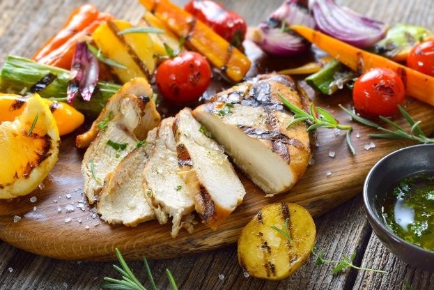 Gegrilltes Haehnchen mit Kraeutern, serviert auf einem Holzbrett, begleitet von buntem Grillgemuese und einer kleinen Schale Sauce.