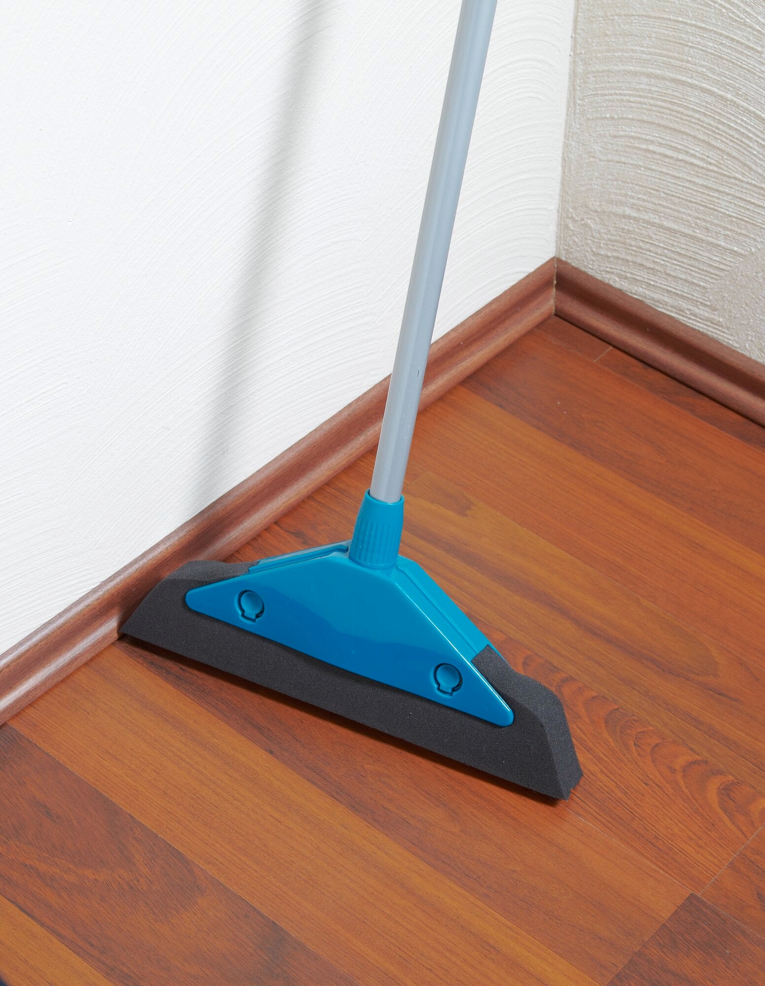 Schuimbezem in actie op een houten vloer. De blauwe bezem met lange steel maakt effectief hoeken en randen schoon, ideaal voor gladde oppervlakken.