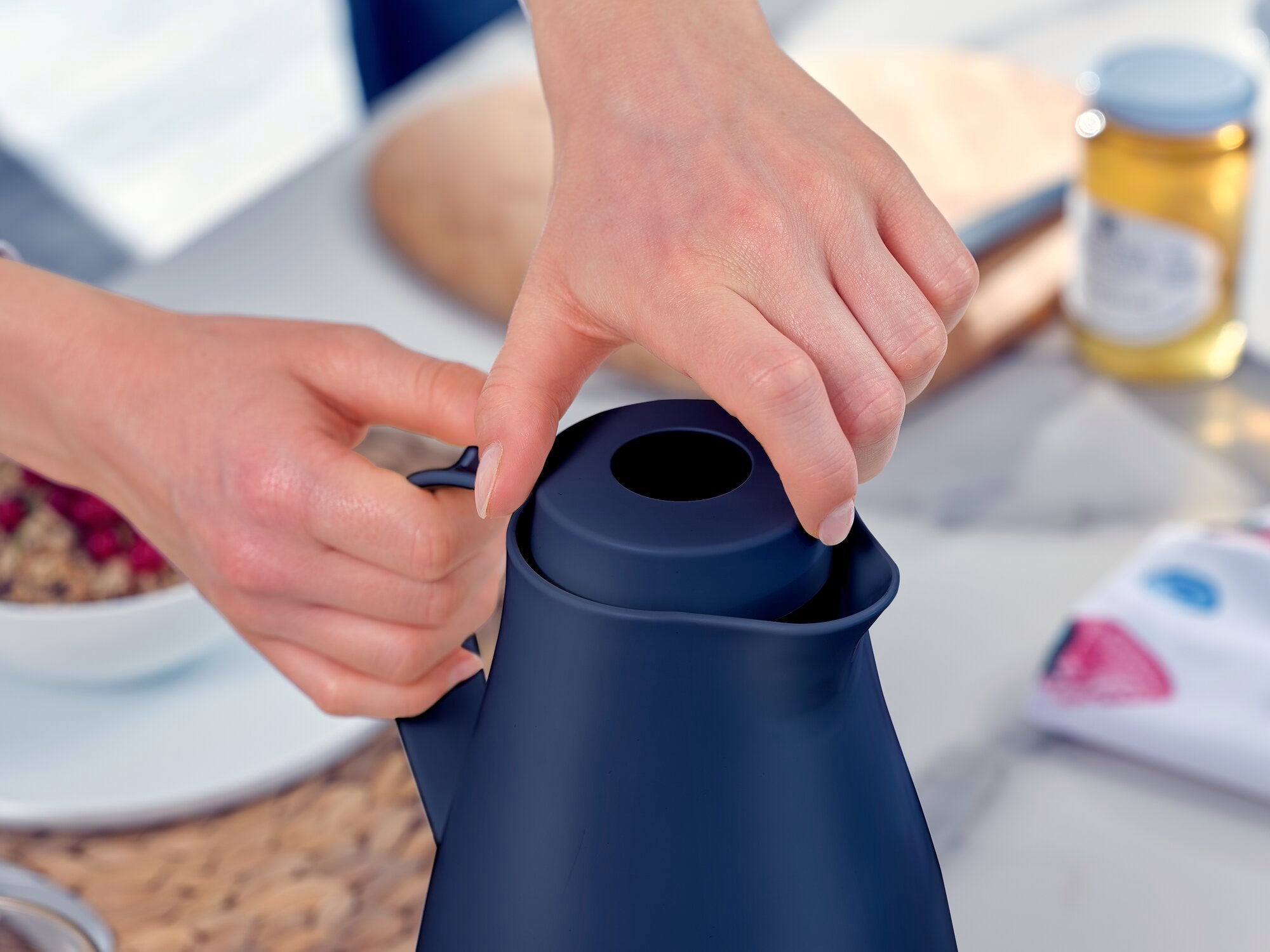Harmonische isoleerkan in gebruik: Een persoon opent het deksel van een donkerblauwe isoleerkan. Op de achtergrond zijn een ontbijttafel en verschillende etenswaren te zien.