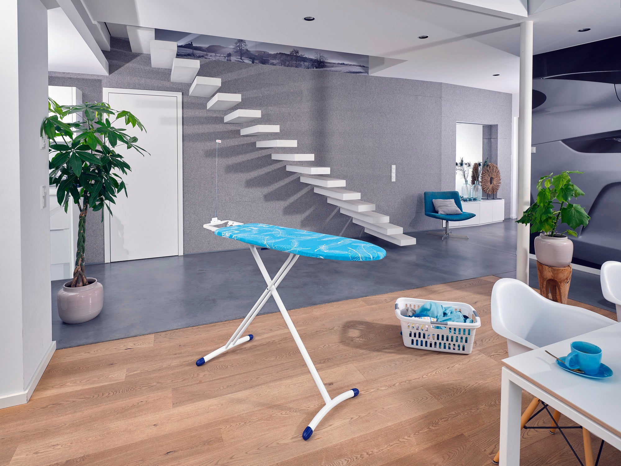Table à repasser Air Board M Solid dans un salon moderne avec parquet. À l'arrière-plan, un escalier, un fauteuil et des plantes. Un panier à linge se trouve à côté.