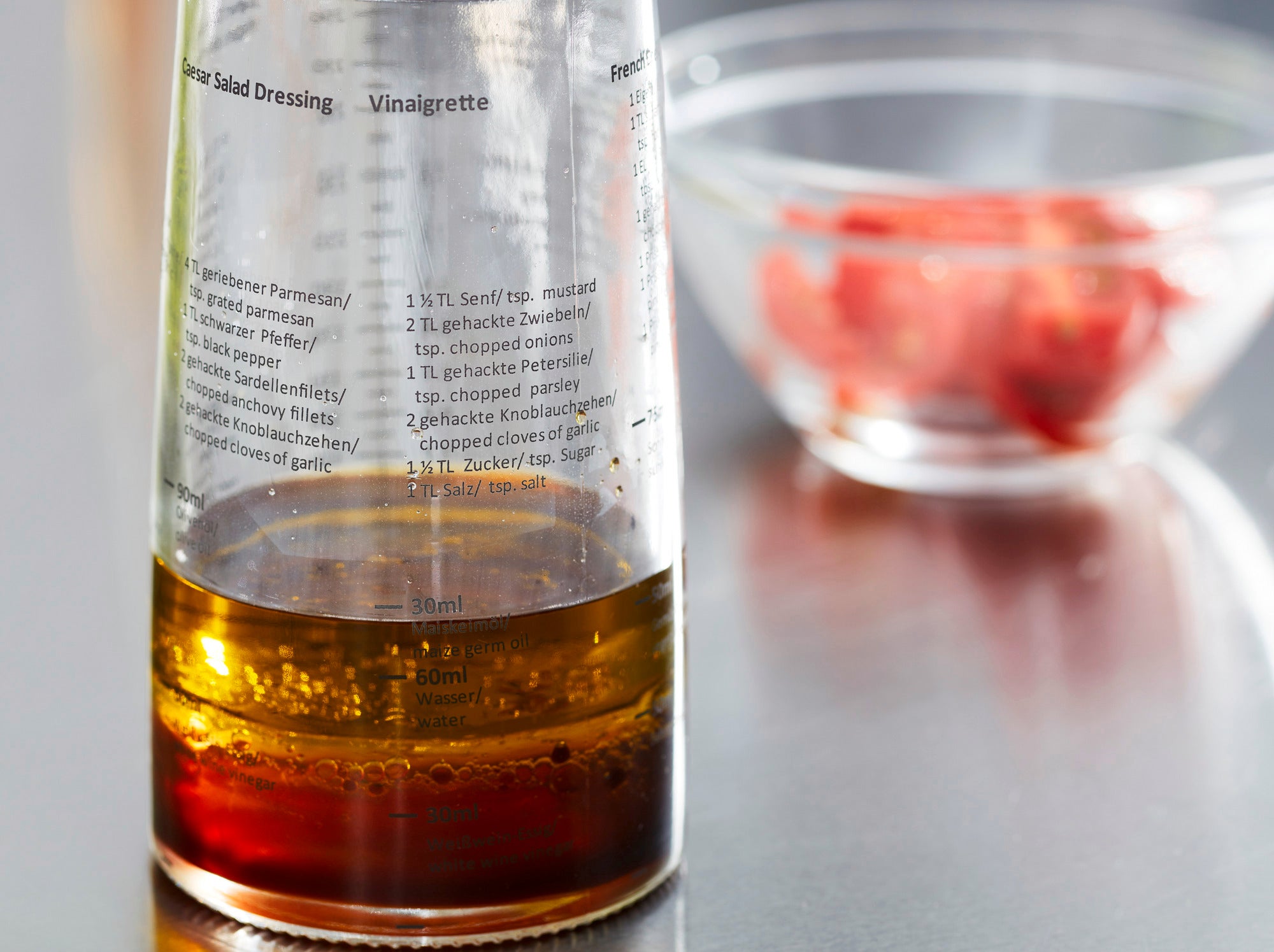 Ein Salat-Dressing-Shaker mit klarer Skala zeigt die Zutaten für eine Vinaigrette. Im Hintergrund ist eine Schüssel mit geschnittenen Tomaten zu sehen. Ideal für die Zubereitung frischer Dressings.