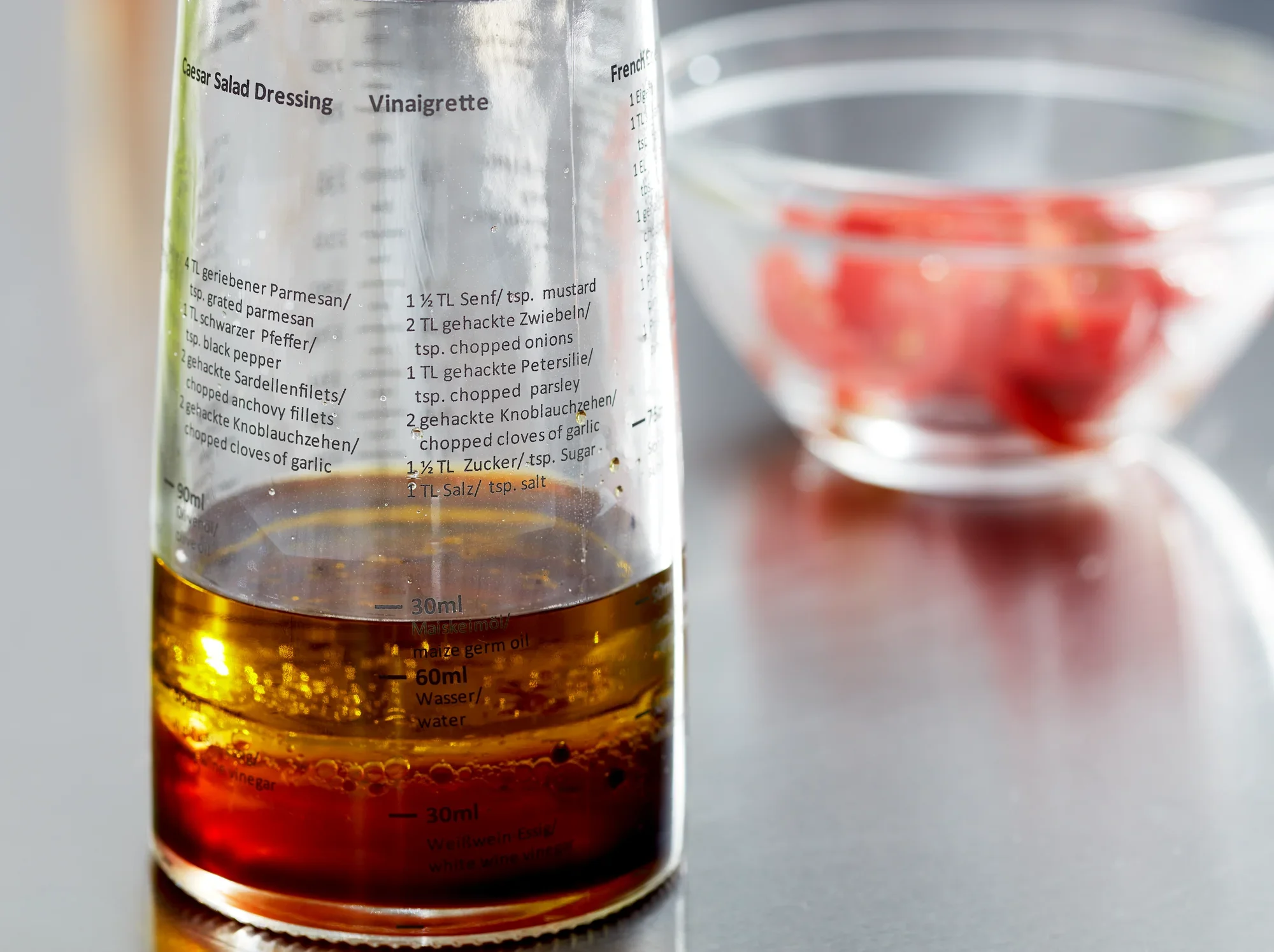 Een saladedressing shaker met een duidelijke schaalverdeling toont de ingrediënten voor een vinaigrette. Op de achtergrond zie je een schaal met gesneden tomaten. Ideaal voor het bereiden van verse dressings.