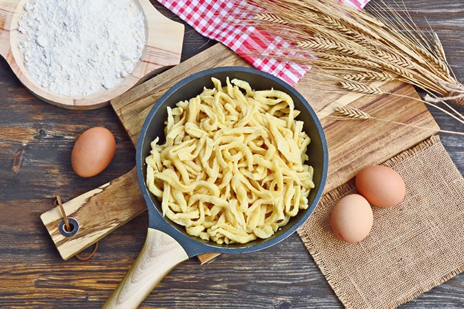 Schnelles Frühlings-Hauptgericht - Kartoffelspätzle mit Geflügel und Rucola Eine Pfanne voller Spaetzle auf einem Holzbrett, umgeben von Eiern, Mehl, Weizenaehren und einem rot-weiss karierten Tuch.