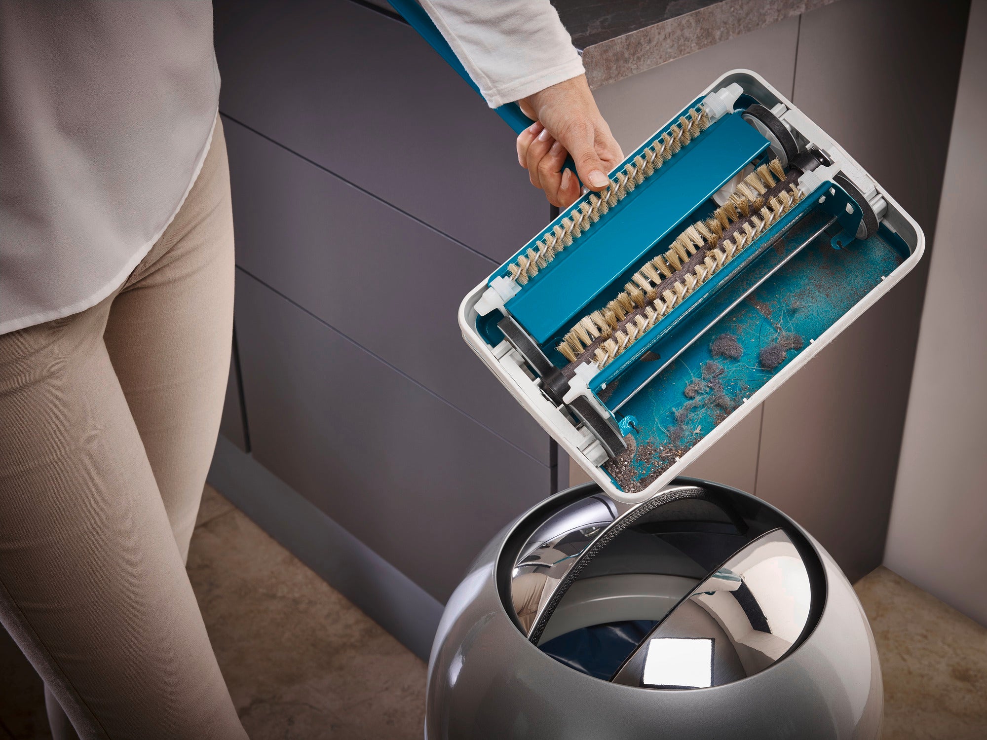 Teppichkehrer Regulus in Türkis wird über einem Mülleimer entleert. Die Bürsten sind sichtbar, während Schmutz und Staub in den Eimer fallen. Ideal für eine gründliche Reinigung.