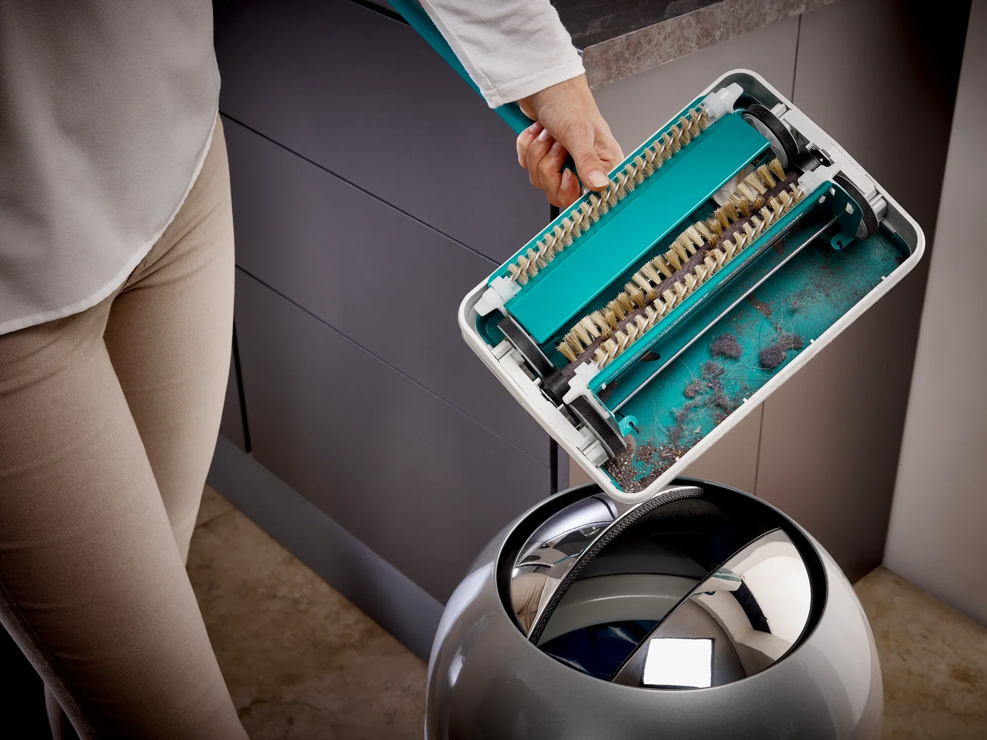 De turquoise Regulus tapijtveger wordt geleegd boven een vuilnisbak. De borstels zijn zichtbaar terwijl vuil en stof in de vuilnisbak vallen. Ideaal voor een grondige reiniging.