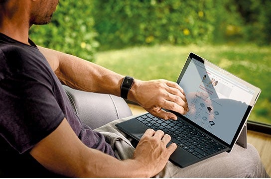 Ein Mann sitzt in Freizeitkleidung auf einem Sessel und bedient ein Laptop, waehrend er aus einem Fenster ins Gruene blickt.