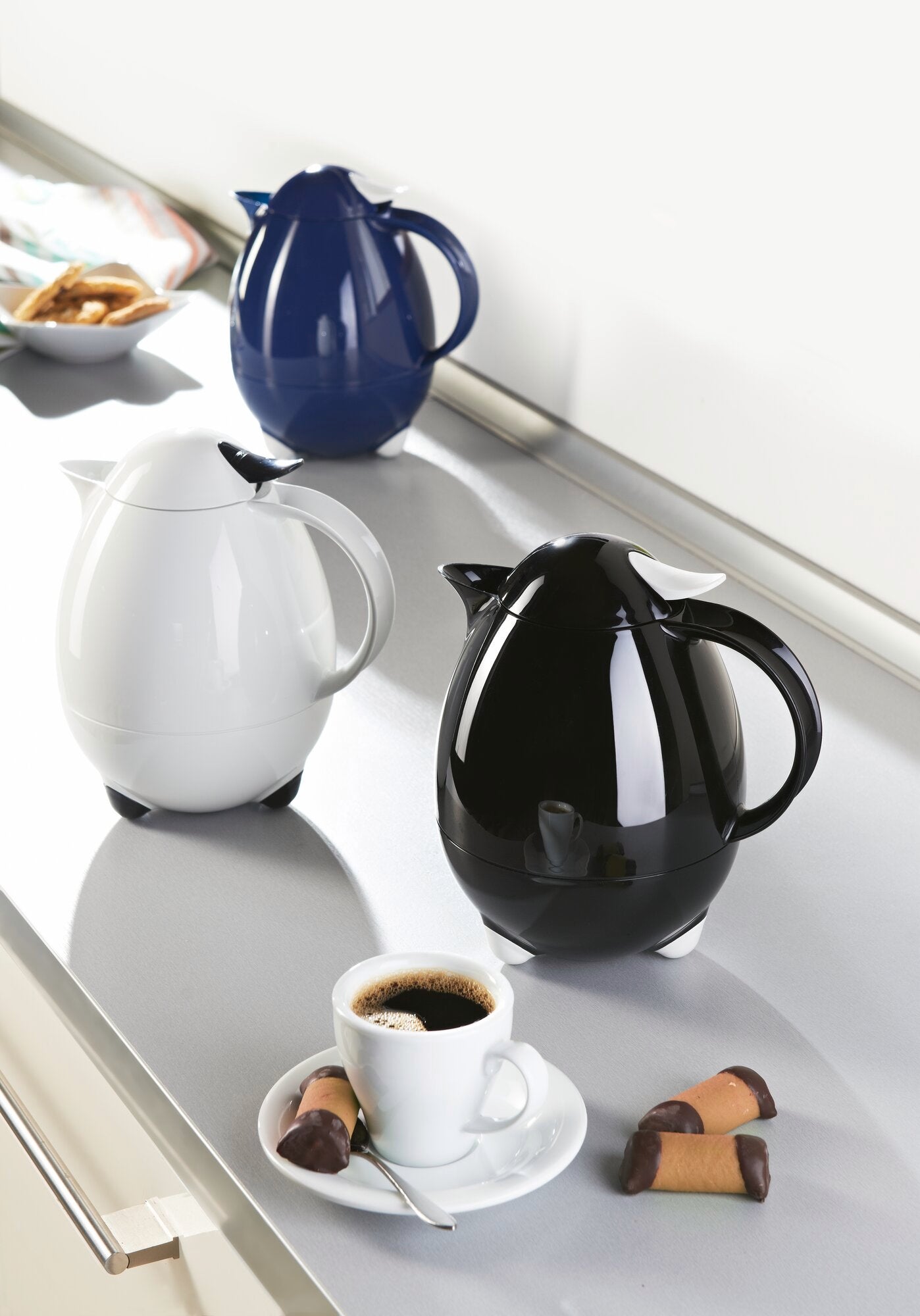 Isolierkannen Columbus in Weiß, Schwarz und Blau auf einer Küchenarbeitsplatte. Im Vordergrund eine weiße Kanne neben einer Tasse Kaffee und Keksen. Ideal für stilvolle Kaffeepausen.