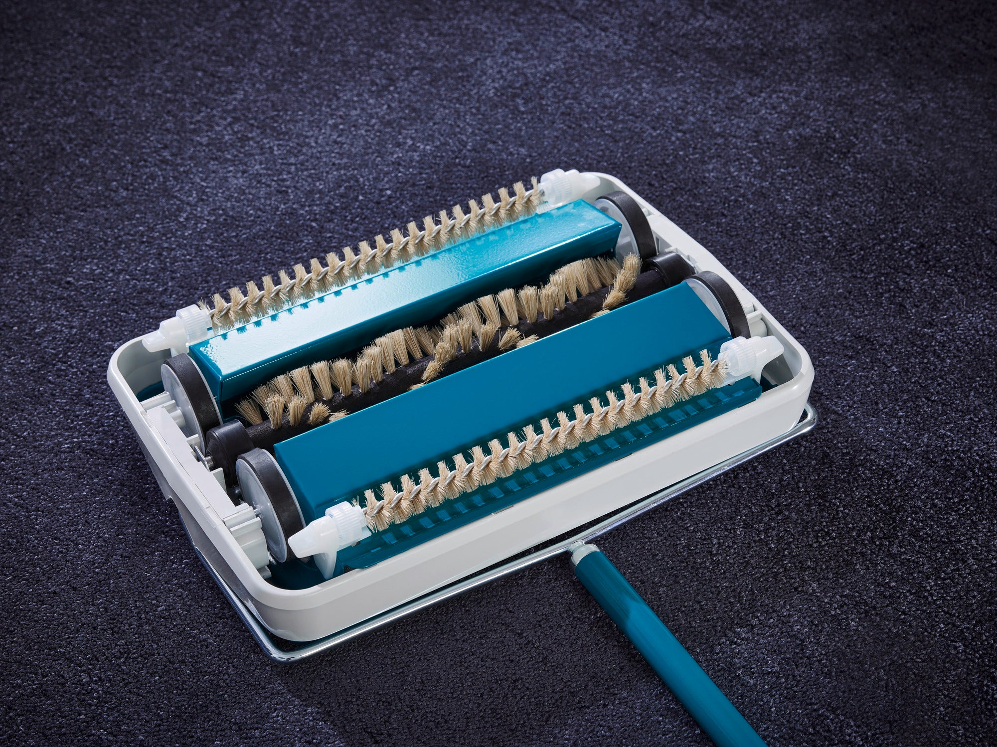 Teppichkehrer Regulus in Türkis auf einem dunklen Teppich. Das Gerät zeigt zwei rotierende Bürsten, die für eine effektive Reinigung sorgen. Ideal für Teppichpflege.