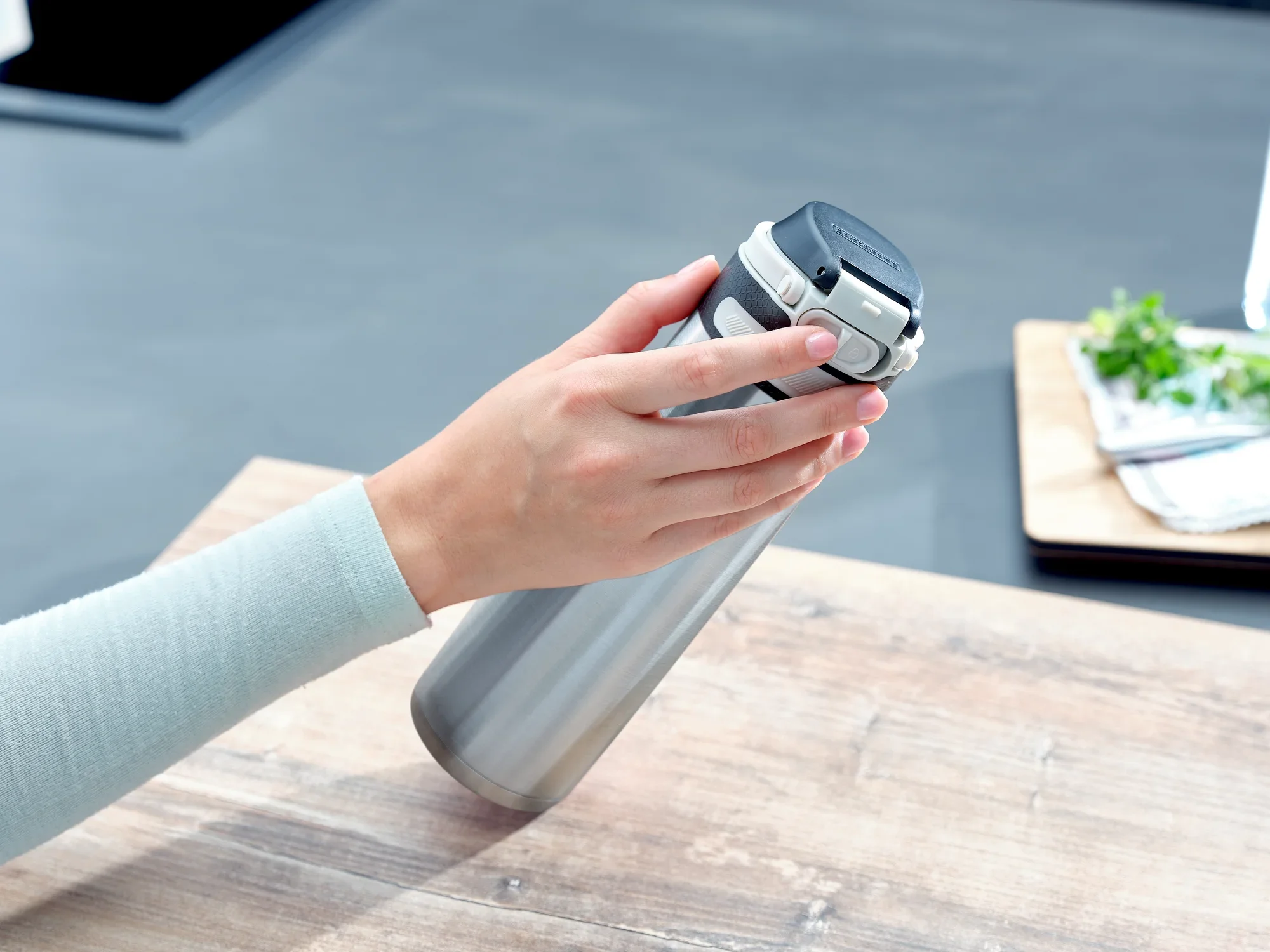 Jedna ręka trzyma srebrny kubek próżniowy Flip o pojemności 600 ml. Kubek ma czarną pokrywkę i leży na drewnianym stole w nowoczesnej kuchni.