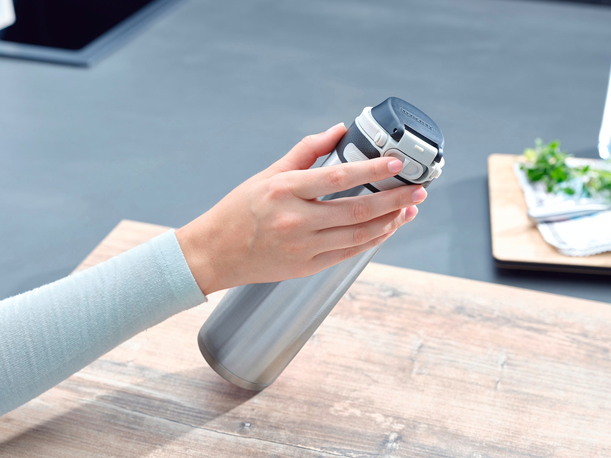 Eén hand houdt een zilveren vacuüm Flip mok vast met een inhoud van 600 ml. De mok heeft een zwart deksel en staat op een houten tafel in een moderne keuken.