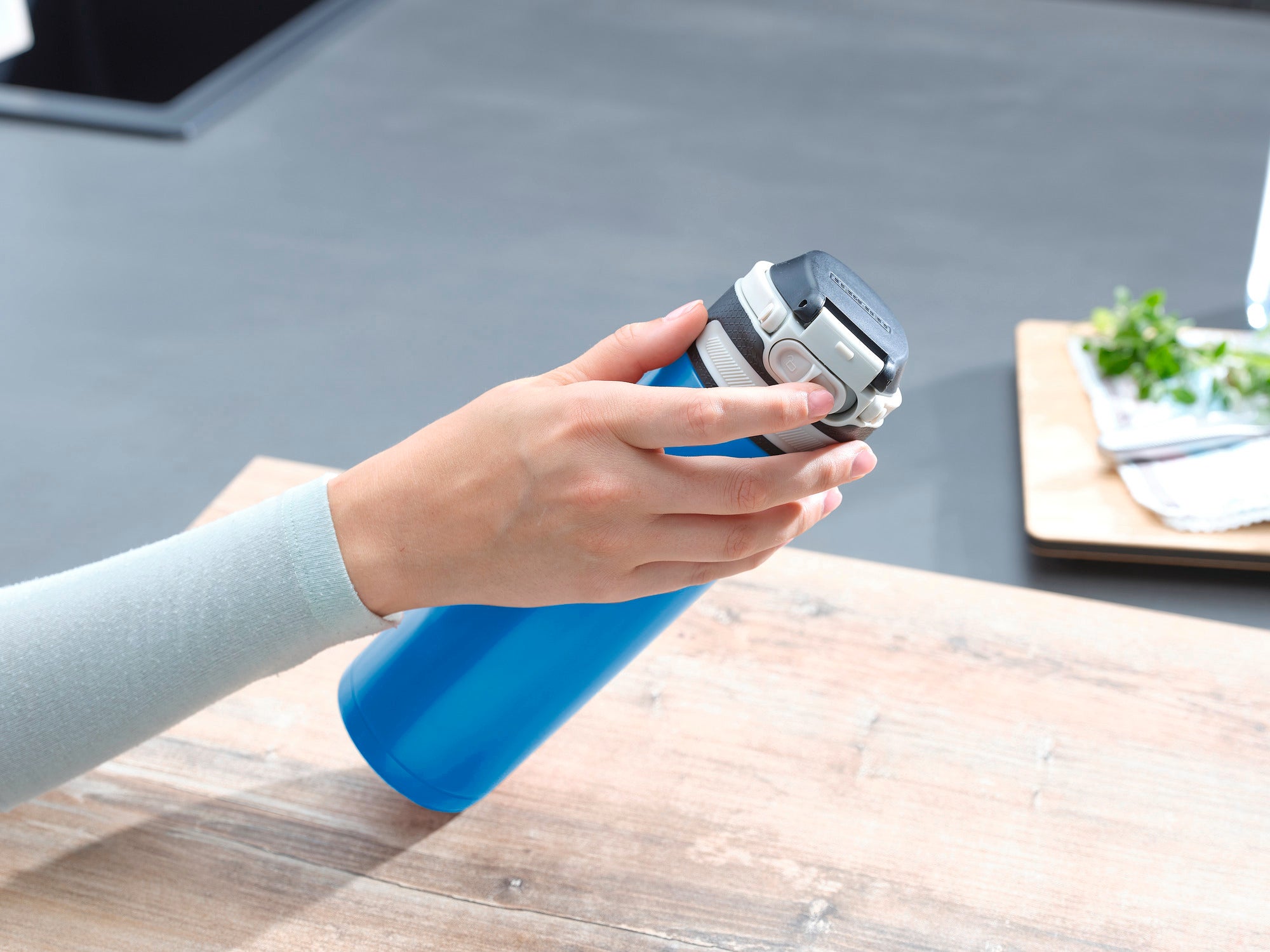 Eine Person hält einen hellblauen Isolierbecher Flip mit 600 ml Fassungsvermögen. Der Becher hat einen praktischen Verschluss und liegt auf einem Holztisch in einer modernen Küche.