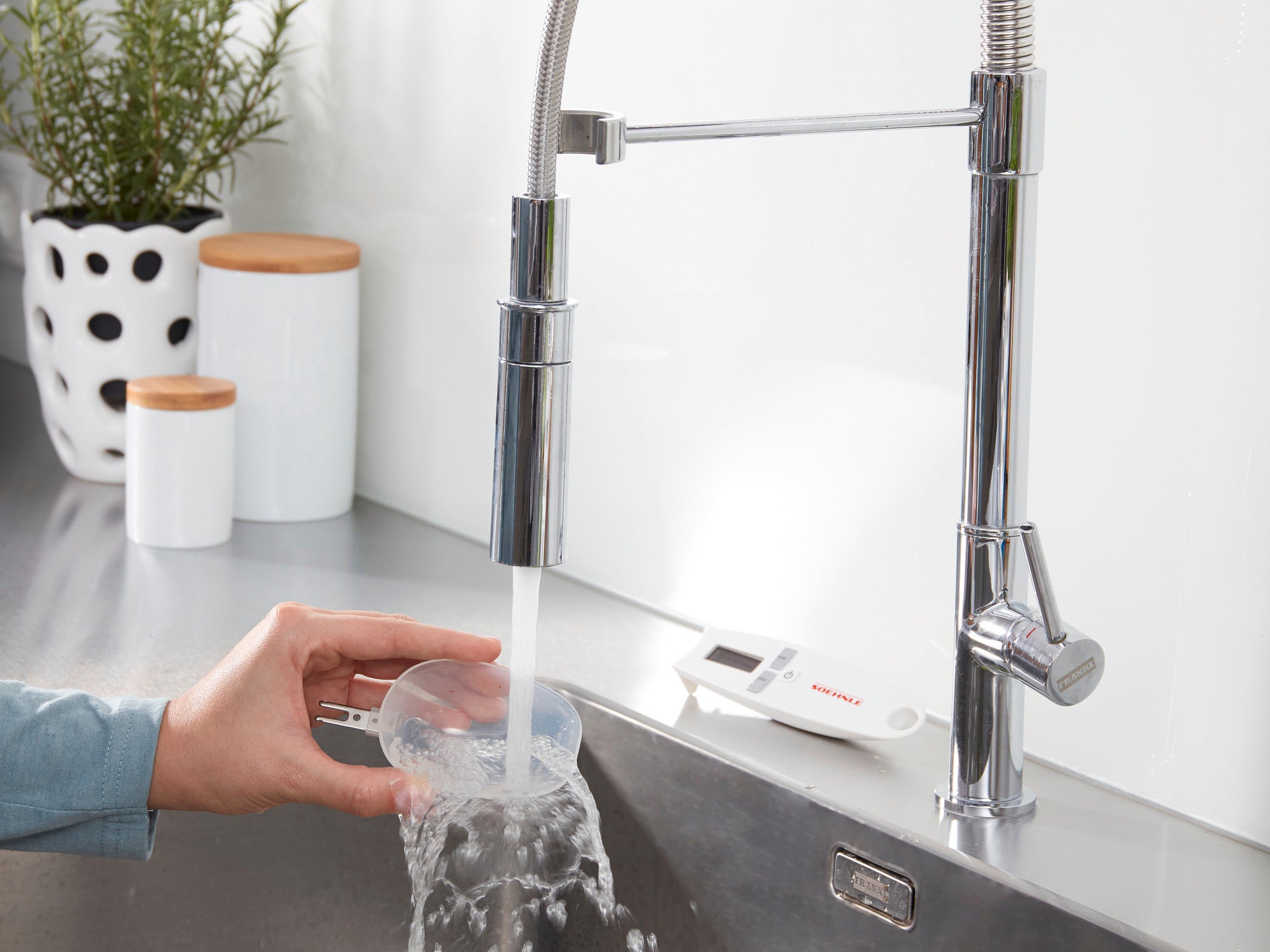 A hand holds a measuring cup under running water in a modern kitchen. The Cooking Star digital kitchen scale can be seen in the background, lying on the worktop.