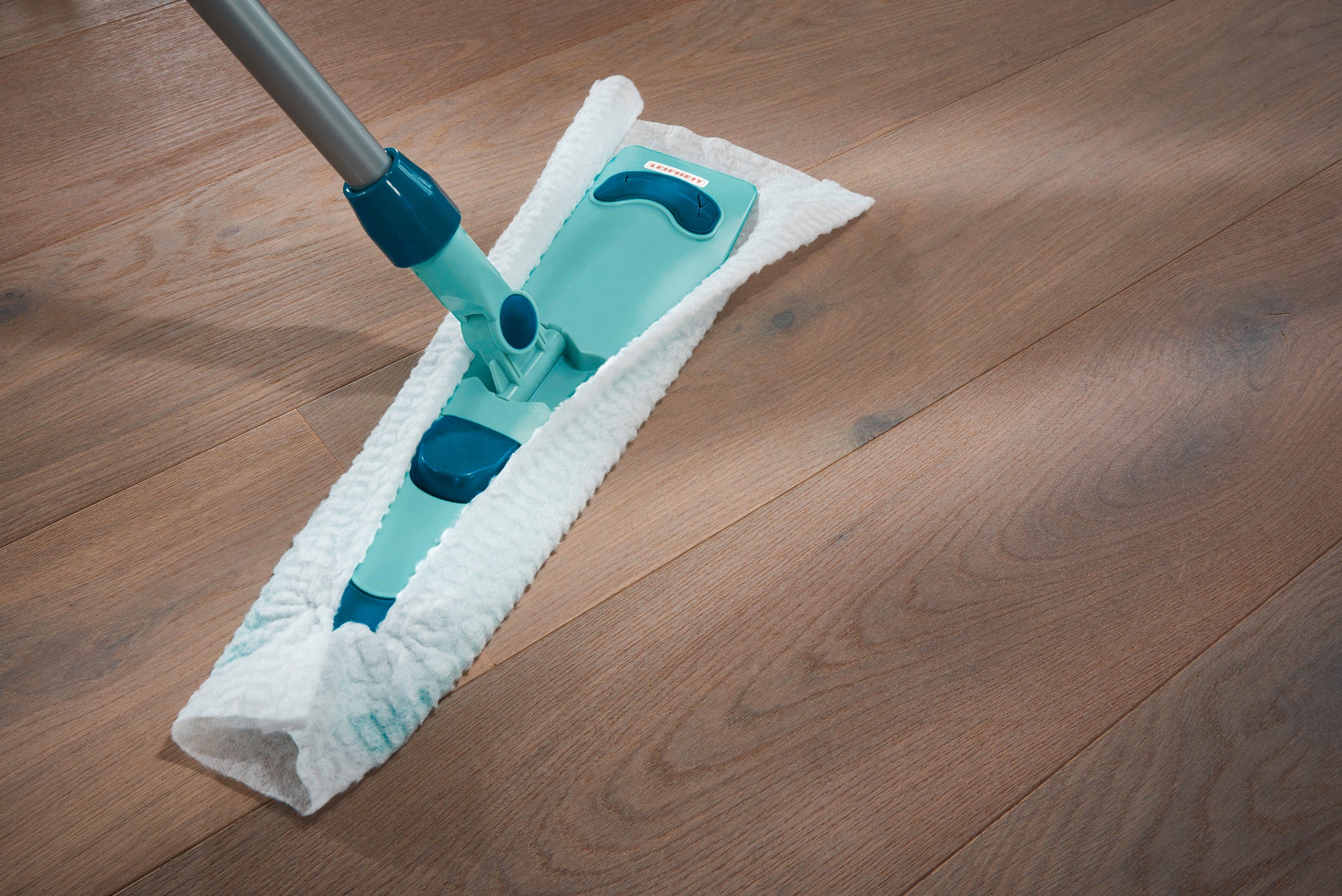 Een vloerwisser maakt een houten vloer schoon. De vloerwisser heeft een blauwe steel en een witte, absorberende doek die vuil effectief verwijdert.