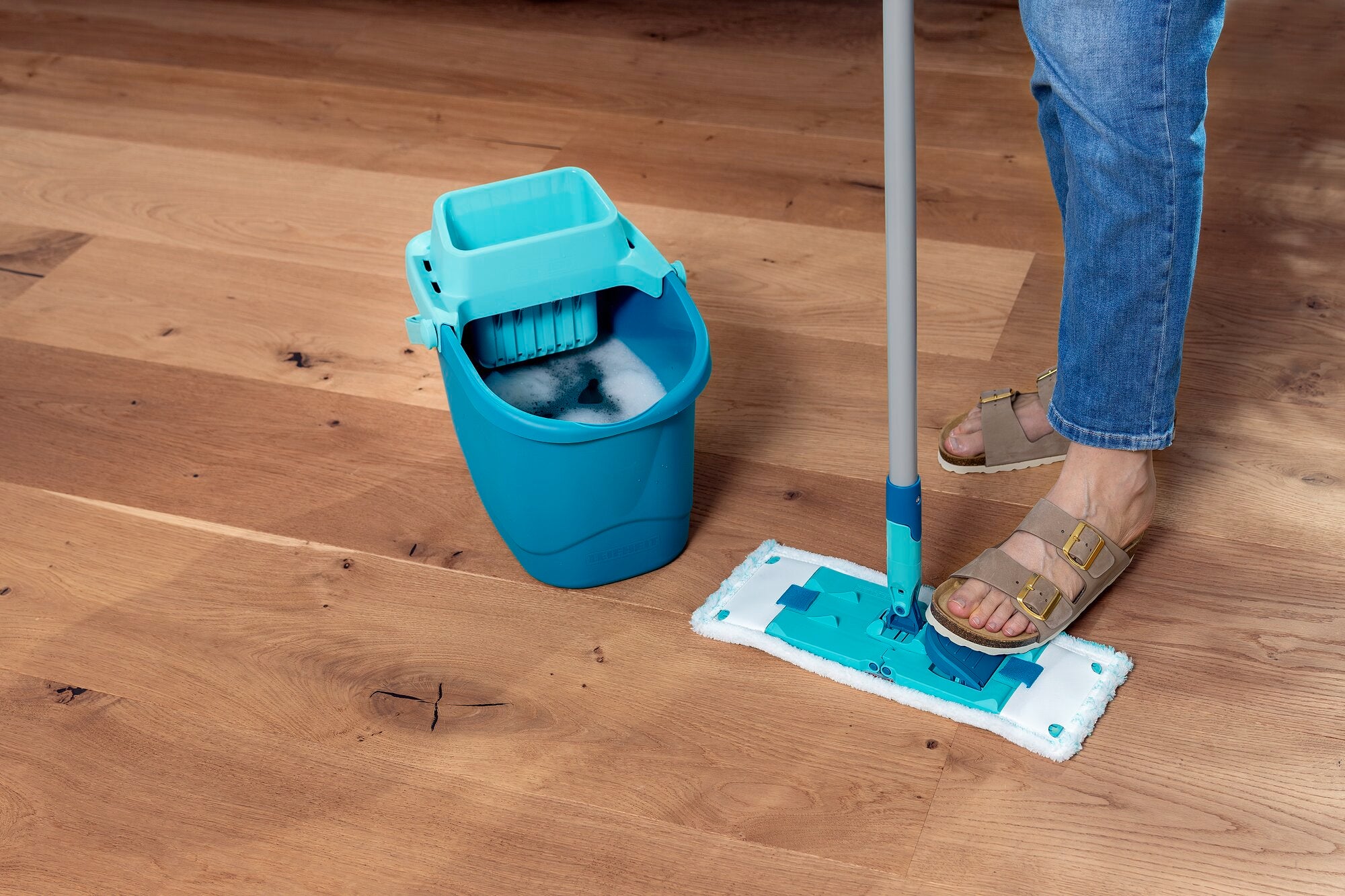 Ein Bodenwischer-Set namens POWER CLEAN wird auf einem Holzboden verwendet. Der Wischer hat ein blaues Design und wird von einer Person mit Sandalen bedient. Ein passender Eimer steht daneben.