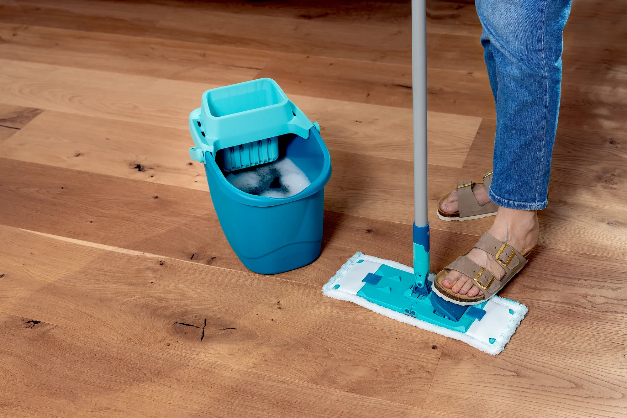 Ein Bodenwischer-Set namens POWER CLEAN wird auf einem Holzboden verwendet. Der Wischer hat ein blaues Design und wird von einer Person mit Sandalen bedient. Ein passender Eimer steht daneben.