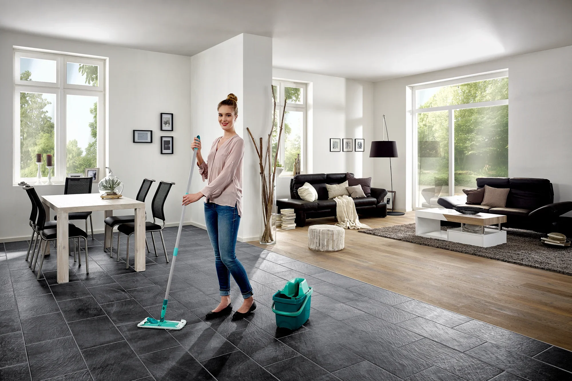 A woman mops the floor of a modern living room with the Combi M floor mop. The room is bright and stylishly furnished with large windows and elegant furniture.