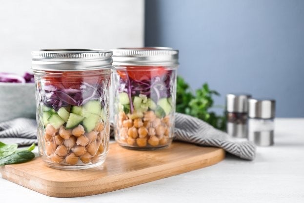 Zwei Einmachglaeser mit geschichteten Kichererbsen, Gurken, Rotkohl und Tomaten stehen auf einem Holzbrett, im Hintergrund befinden sich Kraeutergruen und Salz- und Pfefferstreuer.