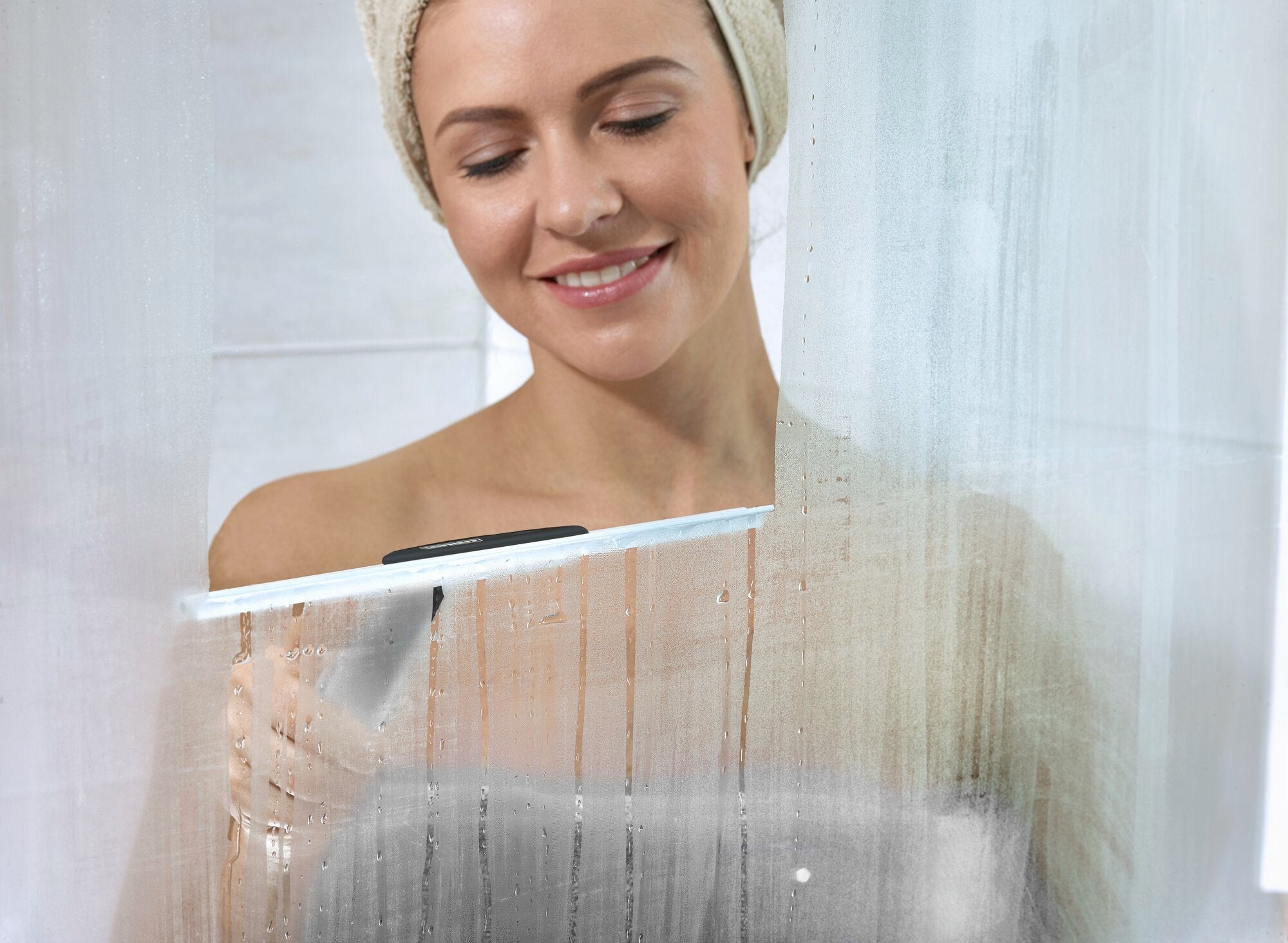 Eine Frau benutzt den Leifheit Duschkabinenabzieher Cabino Black, um eine Glasduschwand zu reinigen. Sie trägt ein Handtuch um den Kopf und lächelt zufrieden, während sie das Wasser abzieht.