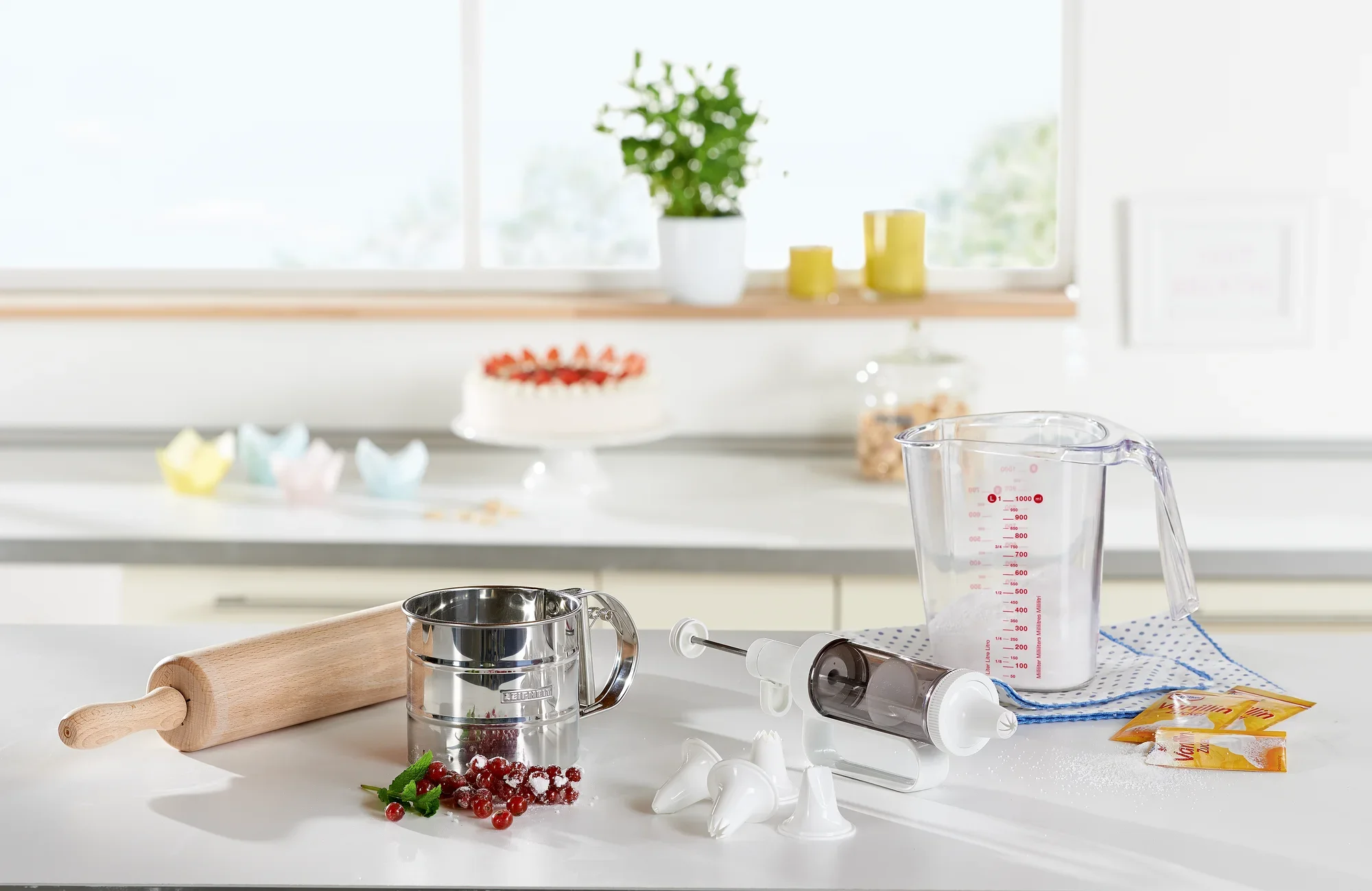 Vaso medidor de 1 l sobre una encimera de cocina con otros utensilios de repostería, como un rodillo, un colador de harina y una manga pastelera. Al fondo se ve una tarta y plantas.