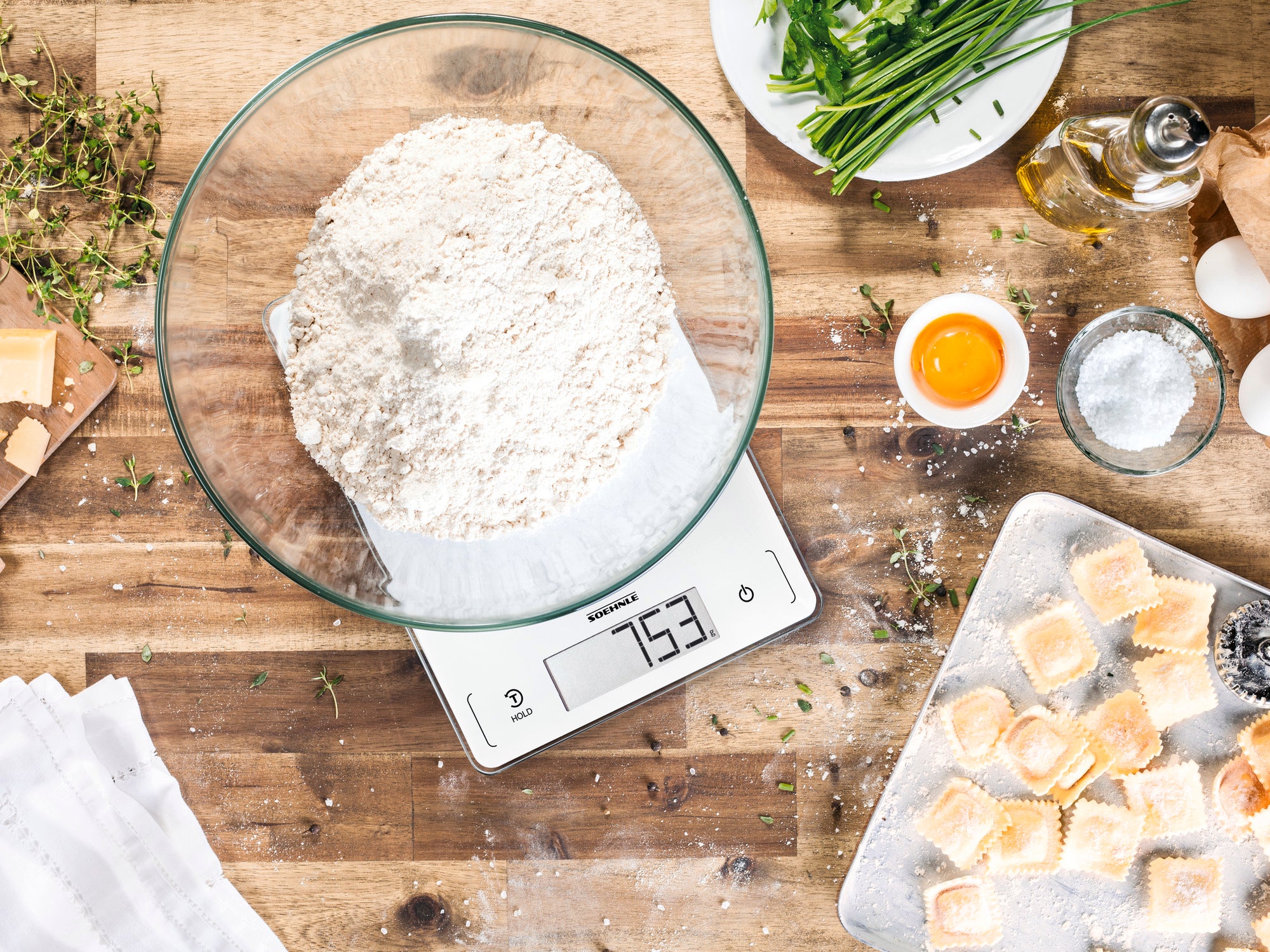 Cyfrowa waga kuchenna wyświetla 753 gramy mąki w szklanej misce. W otoczeniu świeżych ziół, jajek i kawałków ciasta na drewnianej desce, idealnej do pieczenia.