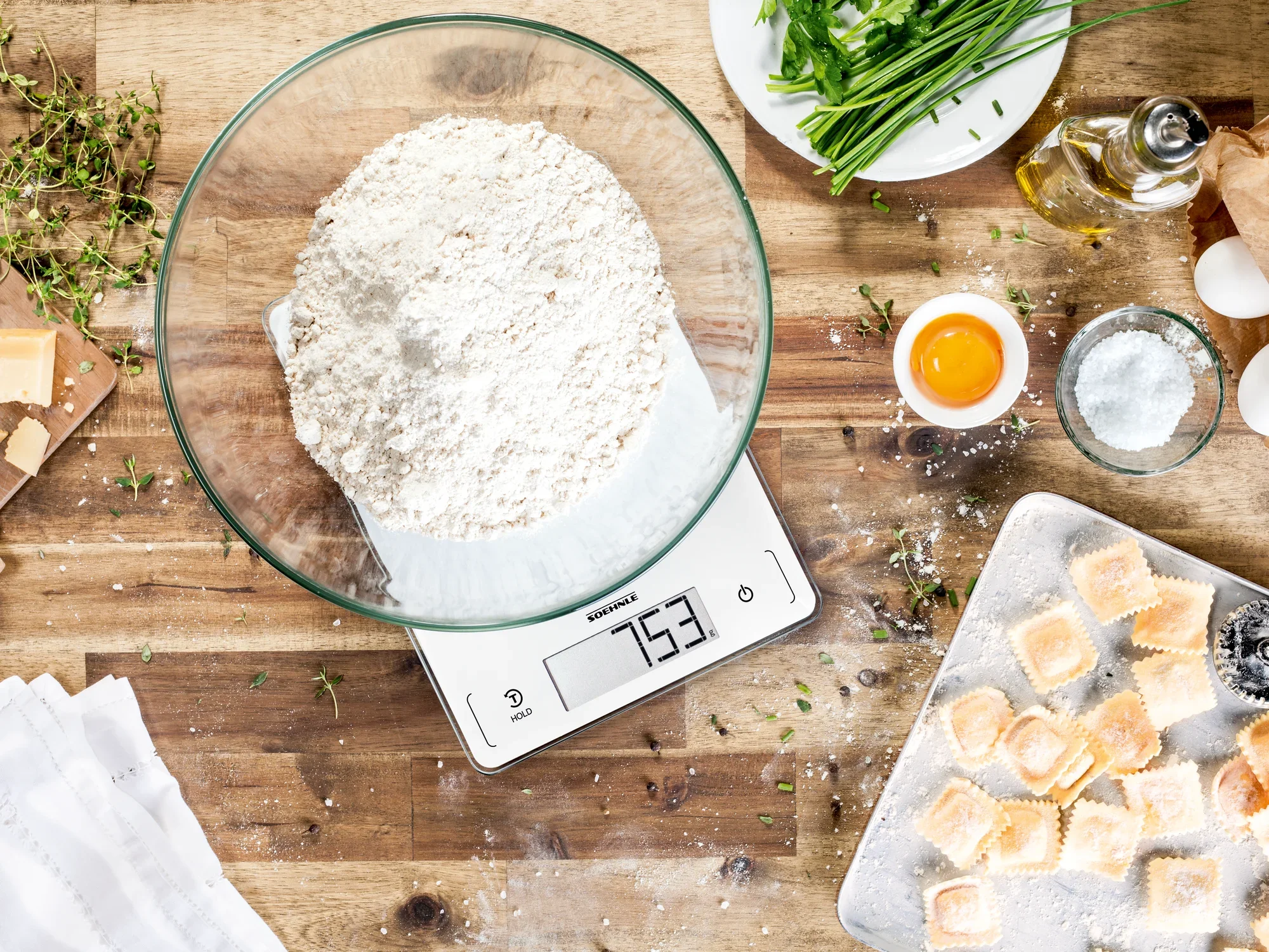 Eine digitale Küchenwaage zeigt 753 Gramm Mehl in einer Glasschüssel an. Umgeben von frischen Kräutern, Eiern und Teigstücken auf einem Holzbrett, ideal zum Backen.