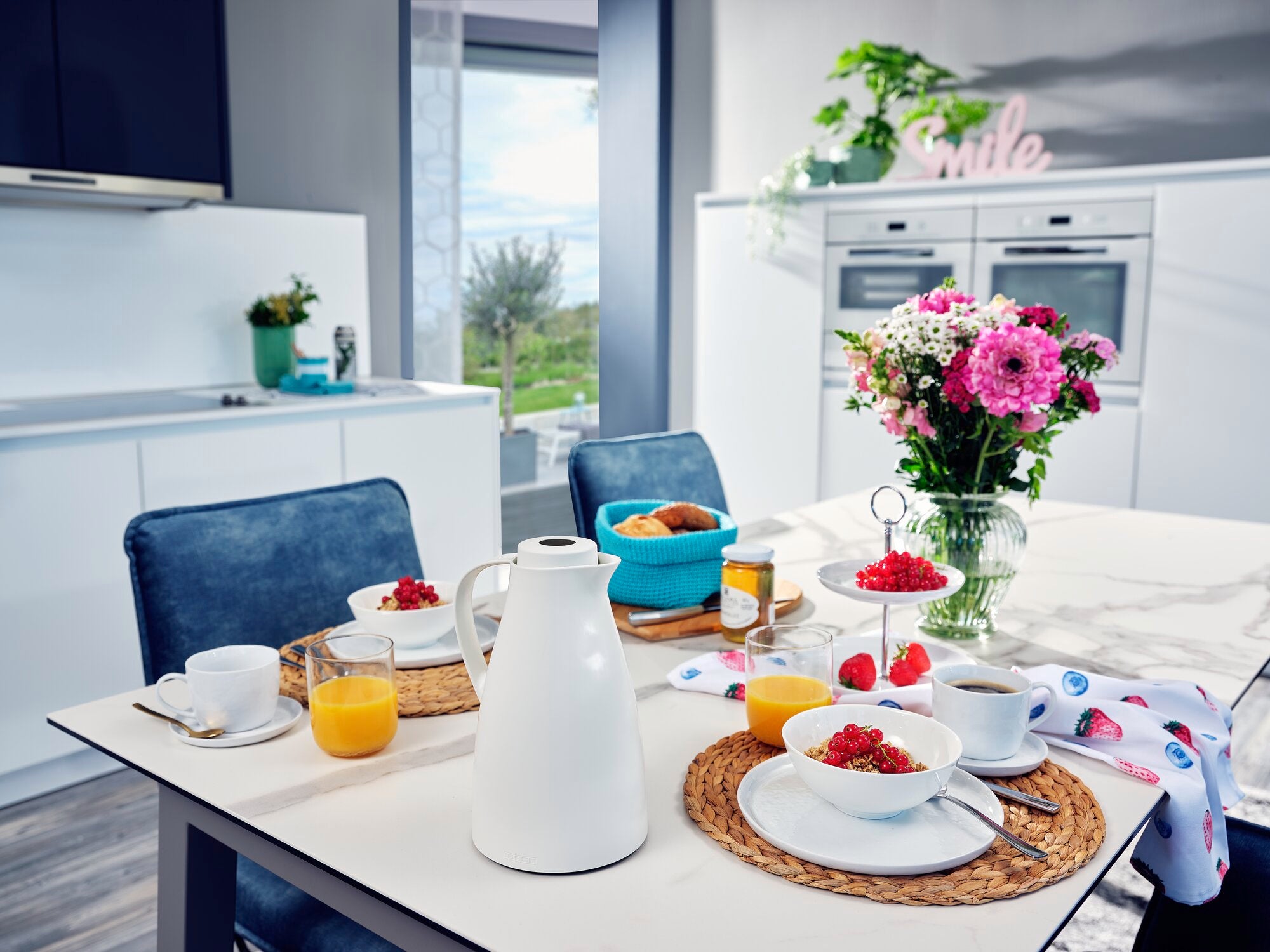 Eine stilvolle Küche mit einer weißen Isolierkanne namens Harmonic auf einem gedeckten Tisch. Umgeben von Frühstückszubehör, Blumen und einem Fenster mit Gartenblick.